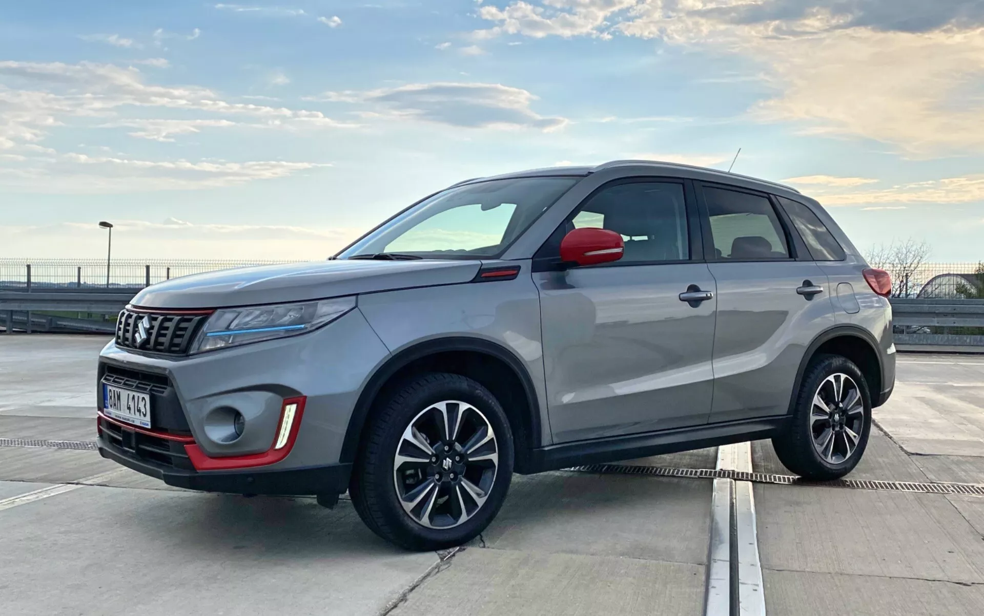 A imagem mostra um Suzuki Vitara 2018, um SUV compacto e moderno, estacionado ao ar livre em condições de luz do dia. É capturada de uma vista frontal de três quartos, destacando tanto o perfil frontal quanto o lateral. O ângulo é ligeiramente baixo, ao nível das maçanetas das portas, sugerindo que o fotógrafo estava agachado ou inclinado. Os detalhes visíveis incluem a grade frontal, faróis, caixa de lâmpadas de neblina, emblema, espelhos laterais, rodas de liga leve com sinais de mudança integrados, maçanetas das portas, aros de janelas, linha do teto e trilhos de teto. Faróis de projetor e antena elegante sugerem tecnologia avançada. Os elementos de design moderno do SUV indicam características como luzes LED e possivelmente um sistema de entrada sem chave.