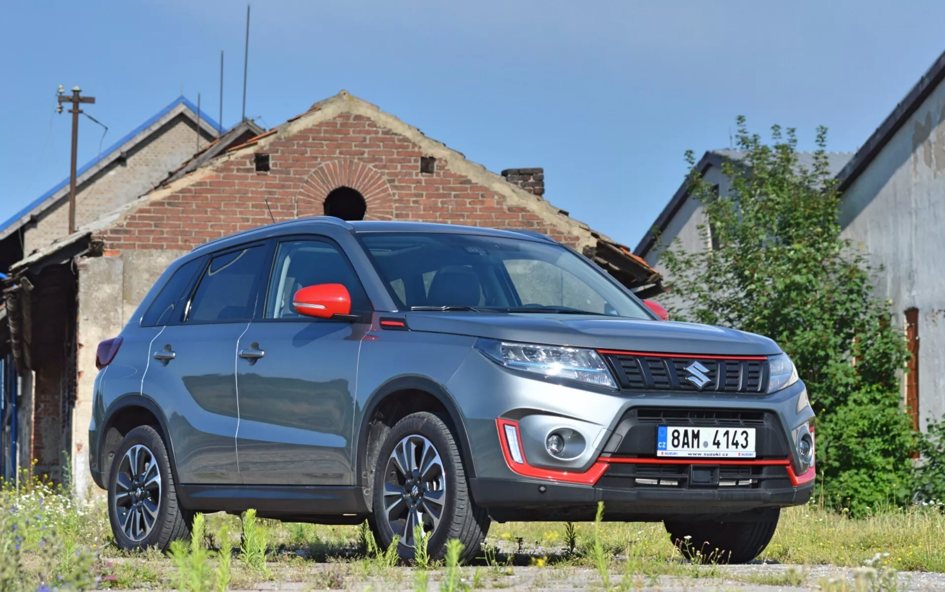 A imagem mostra um SUV Suzuki Vitara 2018 a partir de uma visão frontal de três quartos baixa, destacando o lado direito dianteiro proeminente do veículo com uma notável altura do solo. As características visíveis incluem um para-choque dianteiro robusto com luzes de neblina e chapa de proteção prateada, emblema da marca na grade com ripas horizontais, linhas definidas do capô, faróis modernos possivelmente com tecnologia LED ou HID, rodas de liga leve multisspokes, espelhos laterais vermelhos com sinais de direção integrados, para-lamas musculosos, para-brisa refletor e vidros escurecidos. O Vitara está estacionado em uma superfície de grama não pavimentada, em frente a um prédio antigo que adiciona um fundo industrial ou rural, sugerindo adequação para ambientes externos.