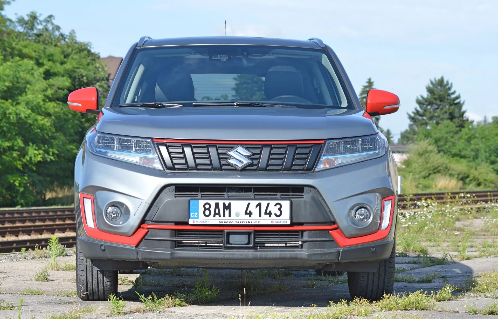 Vista frontal do Suzuki Vitara 2018 a partir de um ângulo ligeiramente baixo. A imagem destaca as características frontais do veículo, incluindo o capô, grade (com emblema Suzuki), faróis elegantes (potencialmente LED) e iluminação adicional no para-choque. O carro parece robusto, sugerindo capacidades off-road. Indicadores de sinalização estão presentes nos espelhos laterais e uma placa dianteira é visível. A carroceria cinza com detalhes em vermelho pode indicar um pacote de design especial. O fundo parece ser uma área industrial abandonada ou um pátio ferroviário, oferecendo uma vista clara do design do carro.