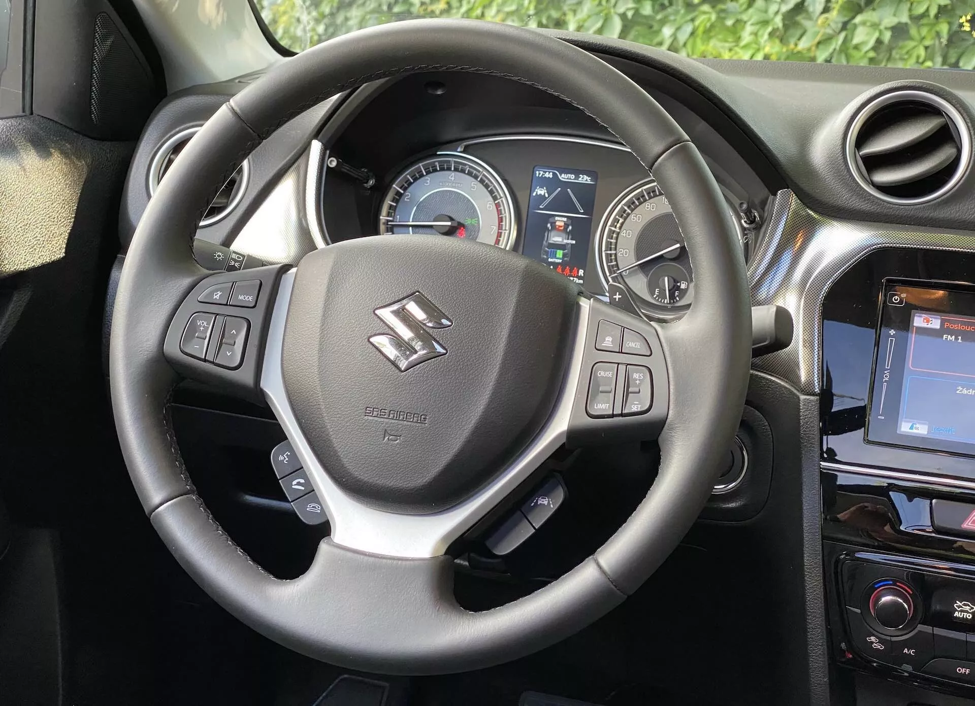 Imagem do interior de um Suzuki Vitara 2018, mostrando a perspectiva do motorista com foco no volante e no painel. O volante, centrado na imagem, apresenta apoios para os polegares e controles para áudio, comandos de voz e controle de cruzeiro. Atrás dele, um painel de instrumentos exibe um velocímetro analógico e parcialmente um visor digital fornecendo informações ao motorista. Uma seção do console central à direita revela um sistema de infotainment com tela sensível ao toque, reforçando a estética moderna do carro.