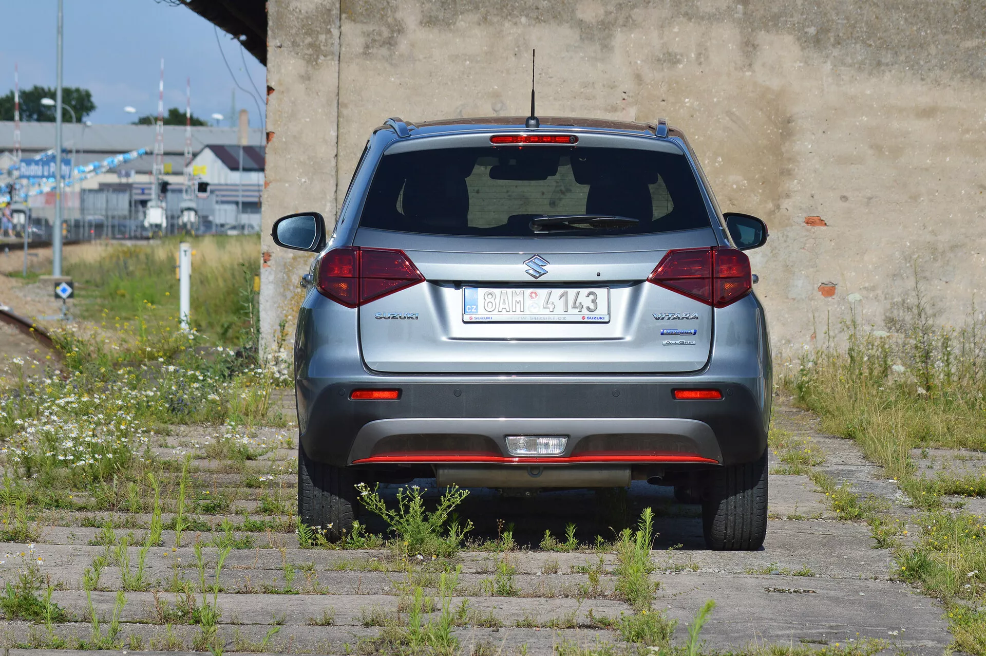 Imagem de ângulo traseiro de um SUV compacto Suzuki Vitara 2018 prateado estacionado em superfície de grama e pavimentação de concreto. A seção traseira completa é visível, com placa central, limpador de para-brisa traseiro, terceira luz de freio, luzes traseiras halógenas padrão, refletores no para-choque, sensores de estacionamento e tubo de escape à esquerda. O veículo está em uma área semi-abandonada com vegetação crescida e uma parede desgastada.
