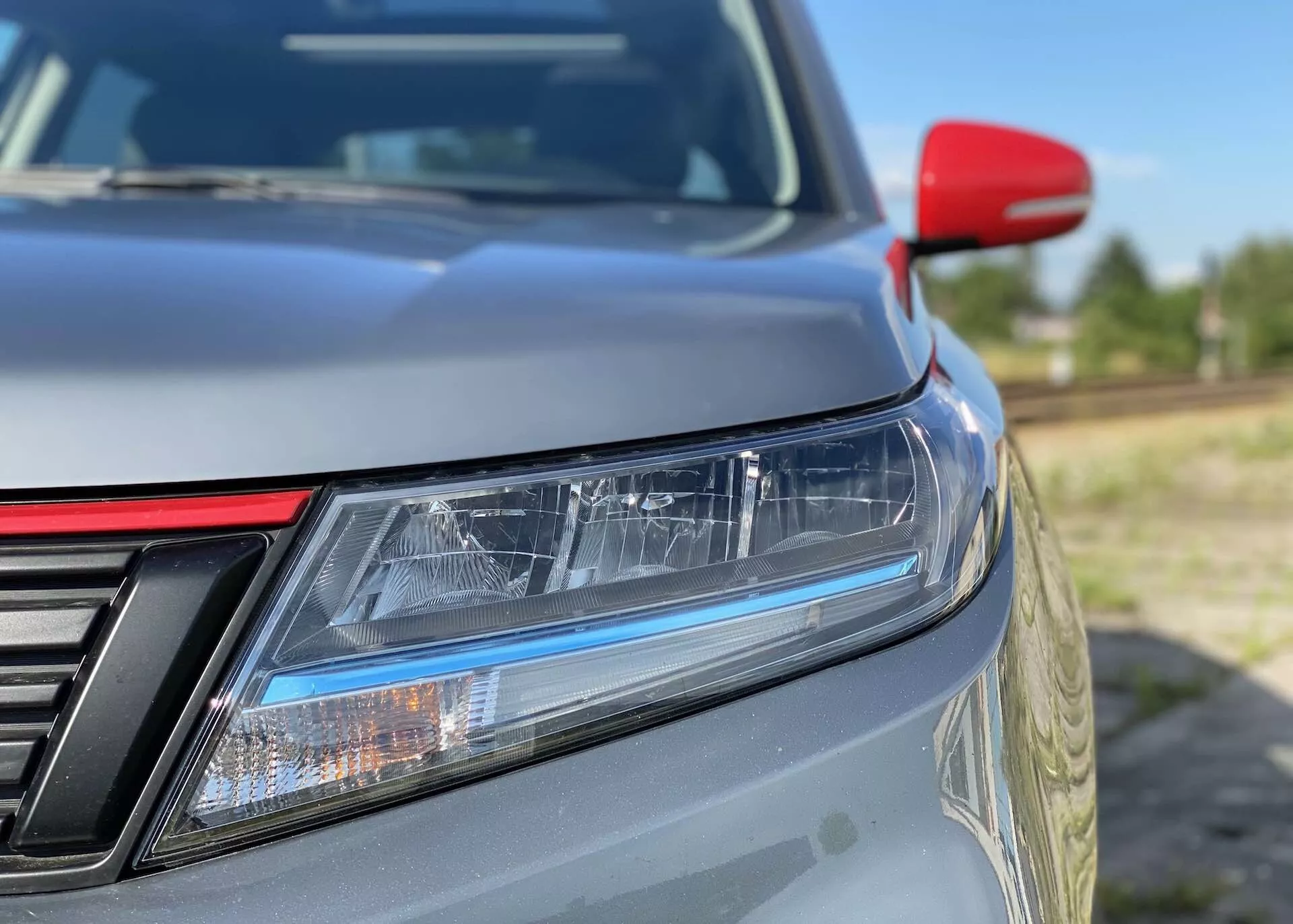 Close-up do canto frontal esquerdo de um Suzuki Vitara 2018, focado em características detalhadas como faróis LED com luzes diurnas, uma grade parcialmente visível com barras verticais, capô com inclinação suave, pintura metálica, um espelho lateral vermelho contrastante possivelmente com indicadores de direção integrados e fundo desfocado que destaca o design elegante e a eficiência aerodinâmica.
