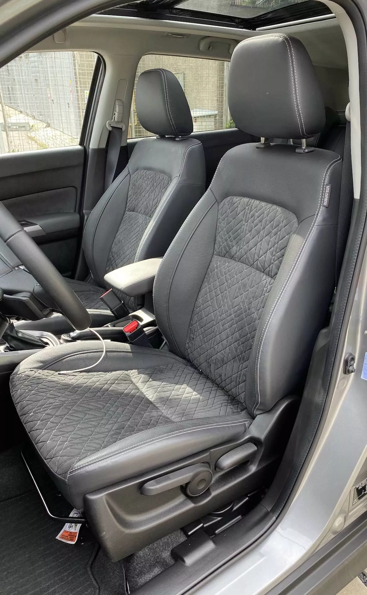 O interior de um Suzuki Vitara 2018, destacando os bancos dianteiros modernos estilo bucket de uma perspectiva ligeiramente acima do nível dos bancos. Os bancos são estofados em couro preto com um padrão acolchoado em losango e reforços laterais em couro liso, oferecendo um equilíbrio entre conforto e esportividade. Uma etiqueta vermelha nos bancos sugere recursos adicionais, como airbags laterais ou bancos aquecidos. É visível o console central com um apoio de braço coberto de couro, bem como um cabo de carregador no banco do passageiro, indicando a presença de portas de carregamento. As fivelas dos cintos de segurança combinam com o esquema de cores preto do interior. Também é visível parte do painel dianteiro e da coluna B, sugerindo uma cabine espaçosa.