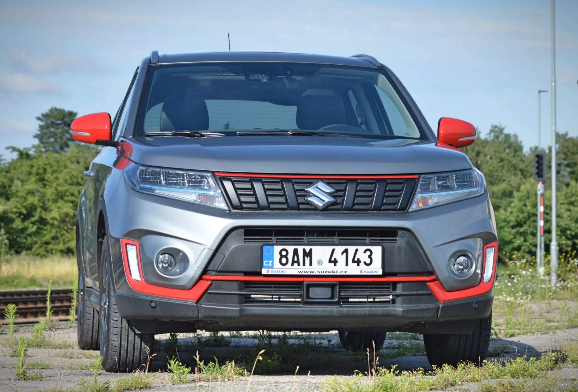 A imagem mostra um moderno SUV Suzuki Vitara 2018 em um leve ângulo frontal, destacando a parte frontal e parte do lado do passageiro contra um fundo de paisagem esparsa com trilhos de trem. O carro, estacionado em uma superfície que parece ser de cascalho, possui uma grade proeminente com design angular, conjunto de faróis elegantes provavelmente com tecnologia LED ou HID, e luzes diurnas ou faróis de neblina integrados no para-choque dianteiro. O para-choque inclui um inserto de cor contrastante que sugere um nível de acabamento off-road ou esportivo, e a altura do solo visível reforça seu caráter de SUV. As linhas elevadas no capô contribuem para um estilo agressivo, enquanto os espelhos laterais e para-lamas mostram um design contemporâneo. As rodas de liga parcialmente visíveis complementam o aspecto robusto. A foto, tirada de perto, permite uma análise detalhada dessas características.