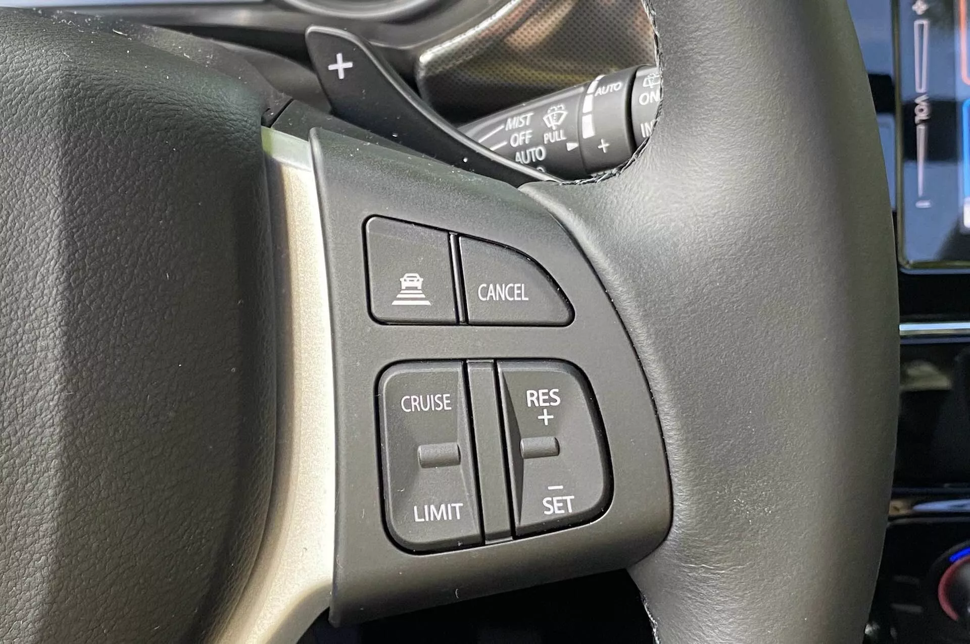 Vista aproximada dos controles montados no volante do Suzuki Vitara 2018, focando no lado esquerdo. O volante em couro preto com costura visível possui botões de fácil acesso, incluindo botão de ativação do controle de cruzeiro, botão de ativação do limitador de velocidade, botões de definição/ajuste do controle de cruzeiro e botão de cancelamento. A imagem também mostra uma haste de controle provavelmente para sinais de direção e faróis, vista da perspectiva do motorista.