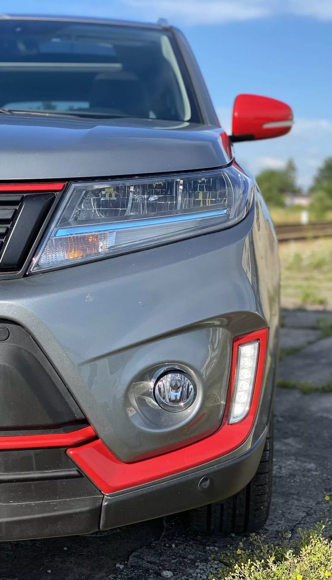 Close-up do quadrante frontal esquerdo do Suzuki Vitara 2018, mostrando faróis LED, luzes diurnas, farol de neblina com acento vermelho, sensores de estacionamento, grelha frontal parcial, espelho lateral com tampa vermelha e pintura cinza metálica.