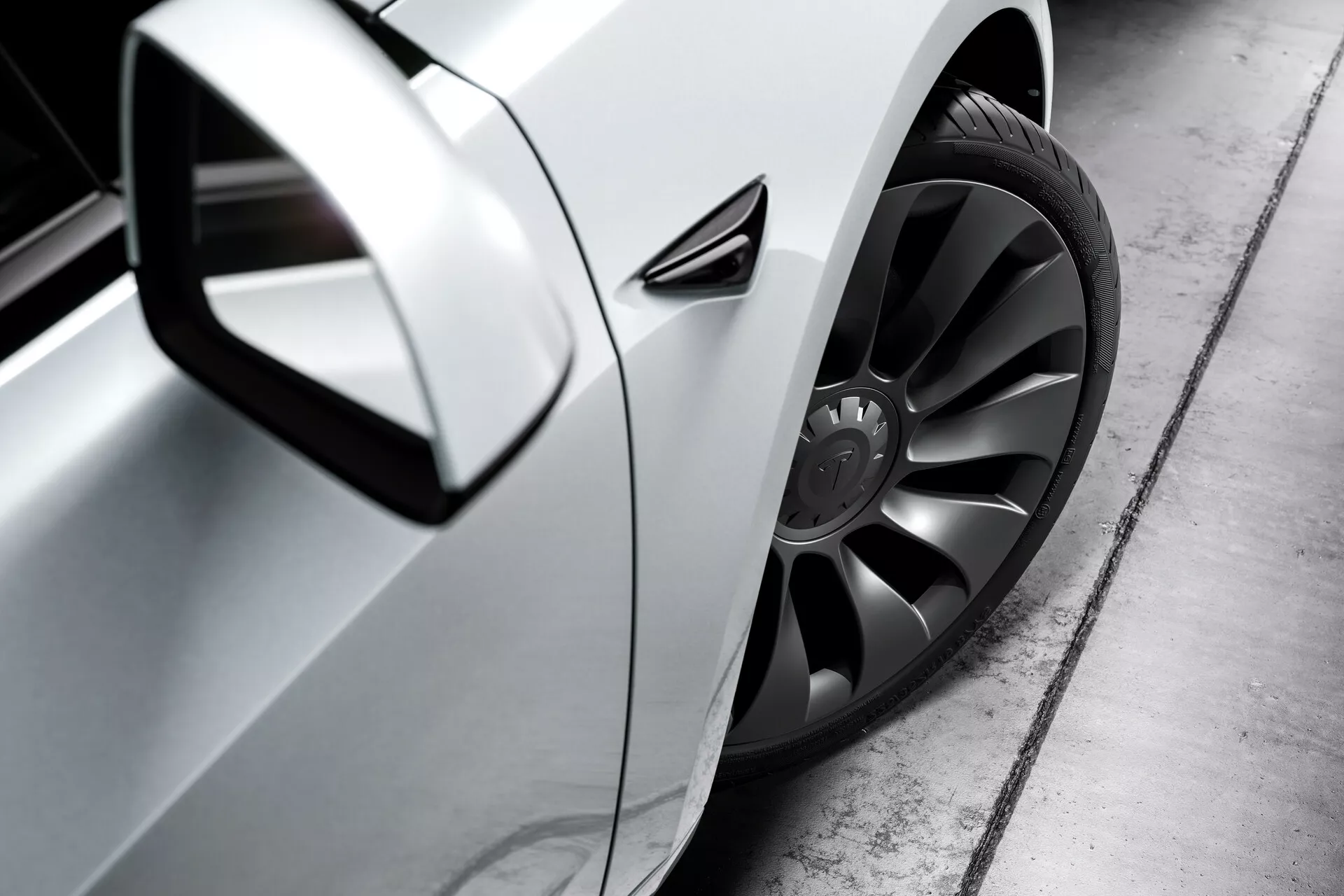 Vista lateral em close de um Tesla Model 3 prateado (2019, 1ª geração) focando na roda dianteira, área da porta do motorista e espelho lateral. A foto, tirada de um ângulo baixo perto do nível do solo, destaca a grande roda de liga leve estilosa com design multirraios escuro e pneu de perfil baixo, painel lateral aerodinâmico com acabamento metálico e espelho lateral aerodinâmico. A maçaneta da porta parece alinhada com a carroceria, sugerindo aerodinâmica avançada, enquanto uma pequena ventilação atrás da roda dianteira pode ser para refrigeração dos freios, aerodinâmica ou estética. O reflexo bem definido no painel indica alta qualidade da pintura.