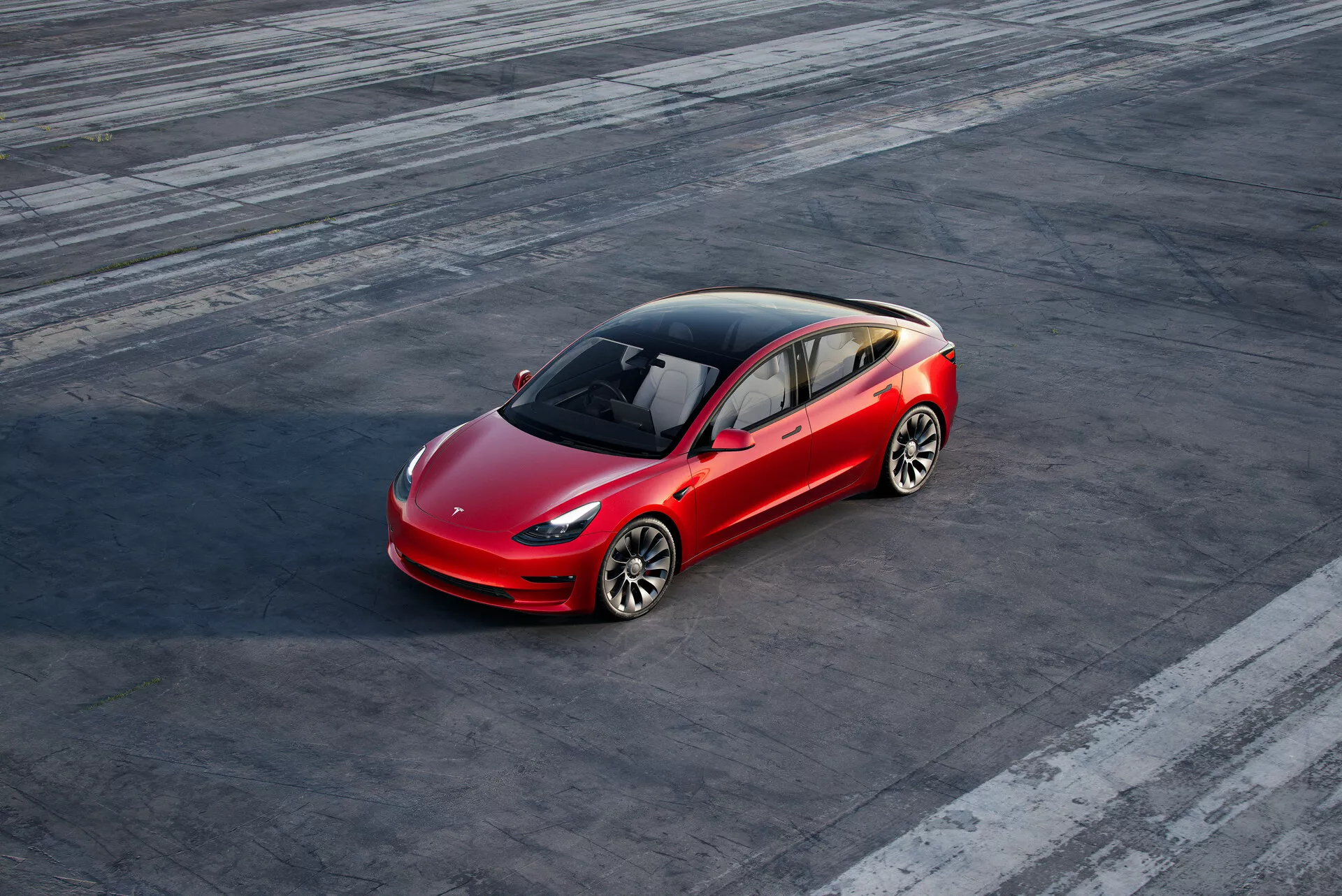 Foto em ângulo alto de um sedan Tesla Model 3 (2019, 1ª geração) vermelho de quatro portas estacionado numa superfície de asfalto texturizado com marcas de pneus visíveis, sugerindo atividade veicular prévia. O carro está ligeiramente posicionado à direita do centro do quadro e é visto de uma perspectiva frontal de três quartos, mostrando o lado do motorista e uma vista parcial traseira. O design aerodinâmico, com uma linha de contorno distinta, teto de vidro panorâmico, rodas de liga leve multirraios, faróis LED e grade dianteira minimalista, destaca a tecnologia avançada e o design elétrico do veículo.