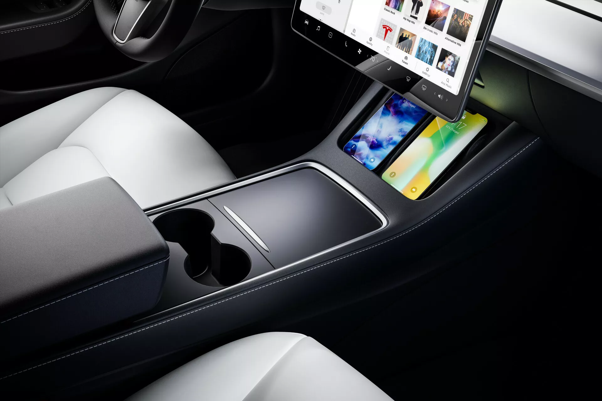 Vista aproximada do interior do Tesla Model 3 2019, destacando o console central e os assentos dianteiros. A imagem, tirada de um ângulo diagonal de cima para baixo, destaca o design elegante e minimalista do console central com uma grande tela sensível ao toque orientada horizontalmente. O sistema de infoentretenimento mostra opções multimídia como integração de smartphone e navegação GPS. Abaixo da tela sensível ao toque há uma superfície plana, dois porta-copos e um compartimento de armazenamento aberto. Os tons escuros do console contrastam com os assentos de couro de cor clara, refletindo um design luxuoso e contemporâneo. No canto inferior esquerdo, é possível ver um comando de controle, adicionando ao ambiente tecnológico do interior moderno do carro.