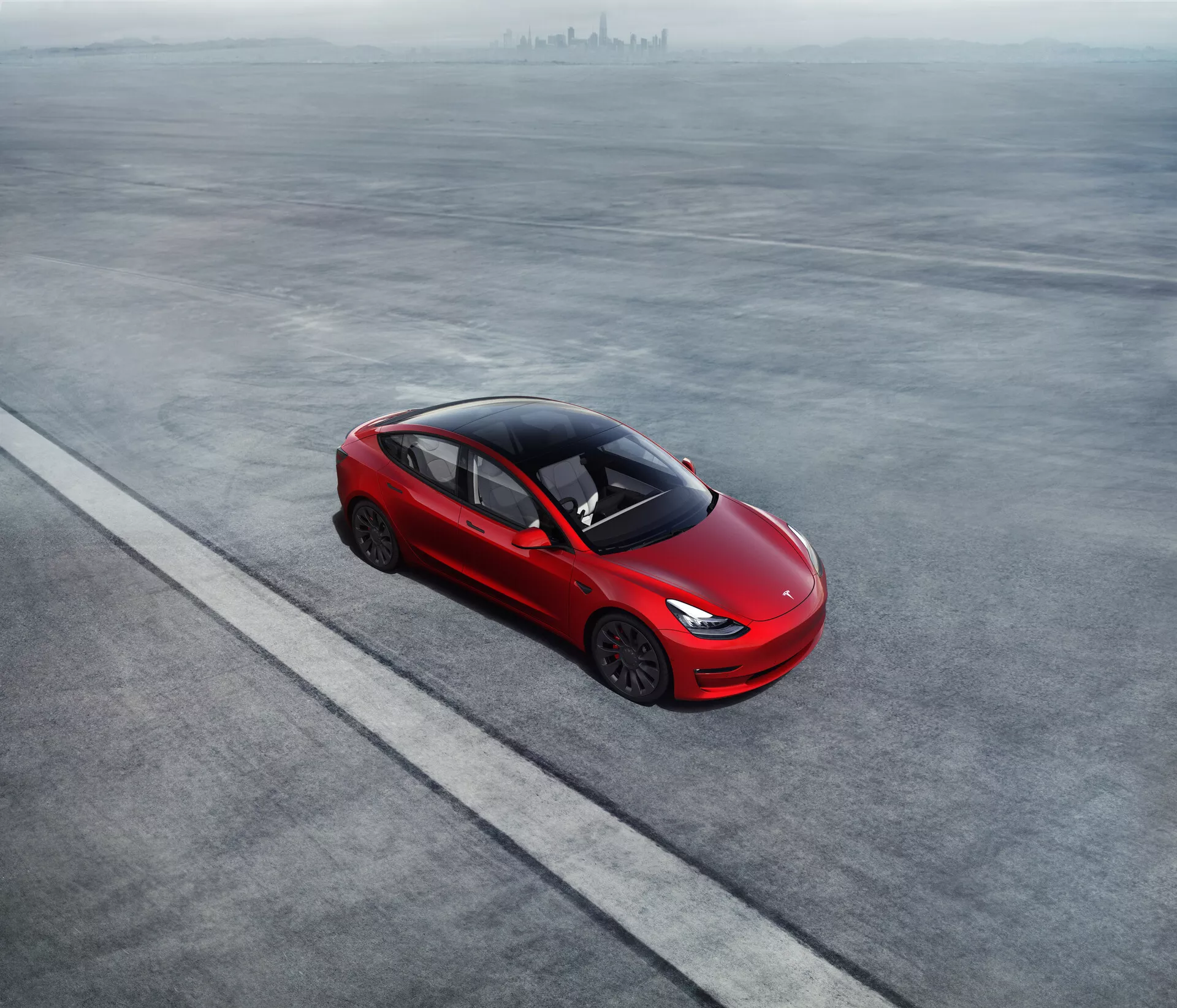 Vista aérea de um Tesla Model 3 2019 (1ª Geração) vermelho intenso estacionado numa superfície plana e cinza. O sedan de quatro portas apresenta uma silhueta aerodinâmica, uma linha de teto inclinada que se funde com o porta-malas e rodas de liga leve únicas. As maçanetas das portas embutidas sugerem eficiência aerodinâmica. O alto grau de tonalidade das janelas, a ausência de escape visível e os faróis finos e modernos implicam tecnologia avançada e possivelmente um trem de força elétrico ou híbrido. O carro está posicionado em paralelo a uma linha reta, enfatizando uma colocação ordenada.