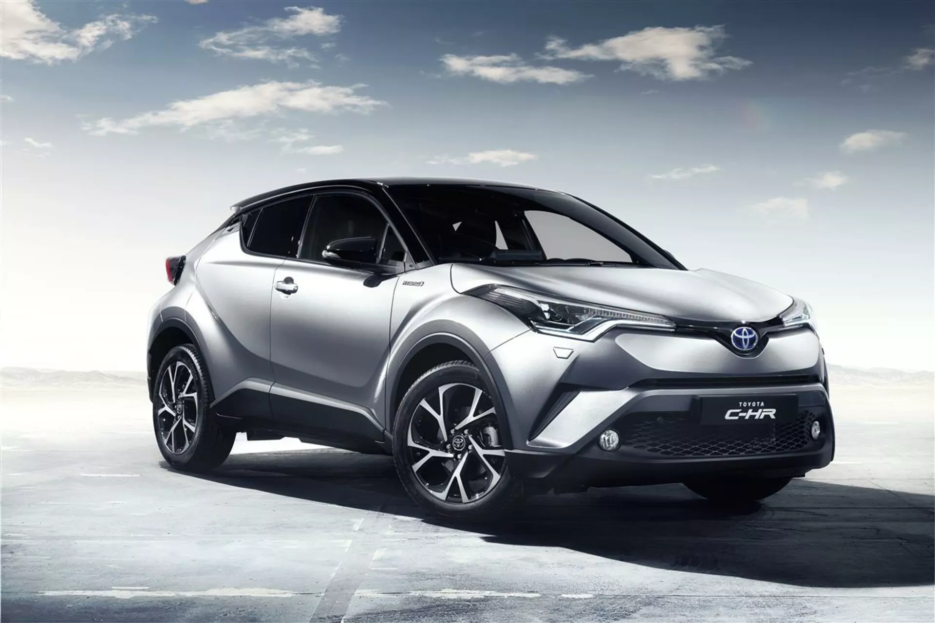 Toyota C-HR 2016 prateado em pose de três quartos frontal mostrando faróis LED, rodas de liga leve e design elegante contra um fundo de céu gerado por computador, destacando suas características esportivas e modernas.
