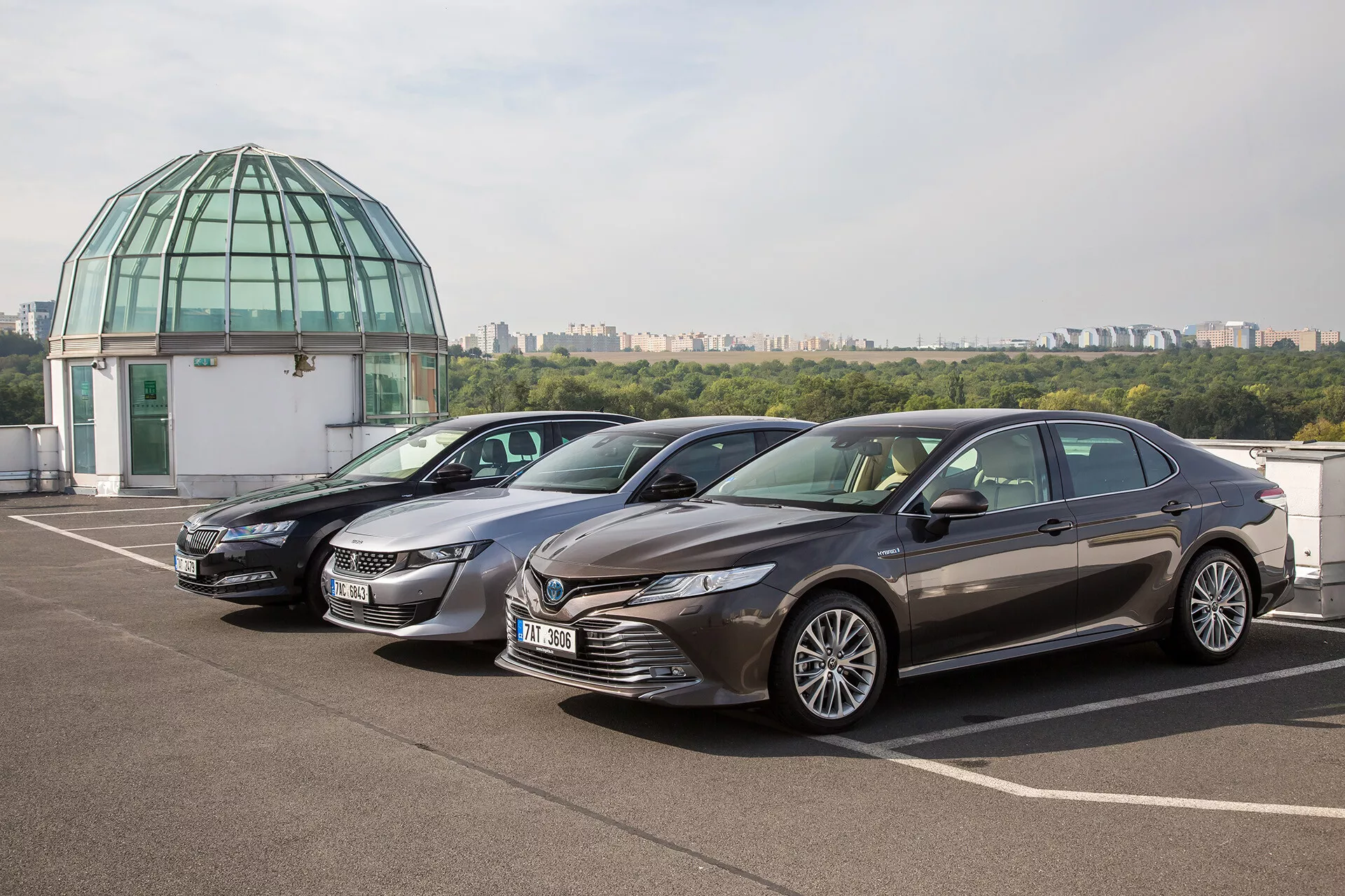 Três sedans Toyota Camry 2019 estacionados lado a lado em um estacionamento no terraço, exibindo seu design elegante e recursos modernos em um cenário urbano diurno com um fundo de cúpula de vidro.