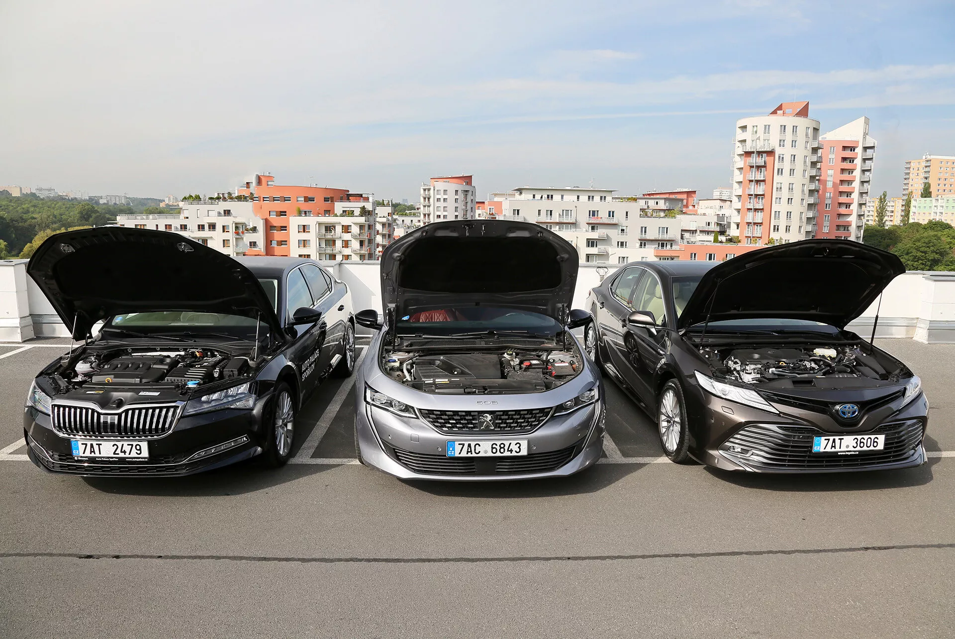 Modelos Limousine Toyota Camry 2019 com capôs abertos mostrando os motores, estacionados lado a lado em uma estrutura elevada, com um horizonte da cidade ao fundo, destacando o design elegante, moderno dos veículos e a tecnologia avançada do motor.
