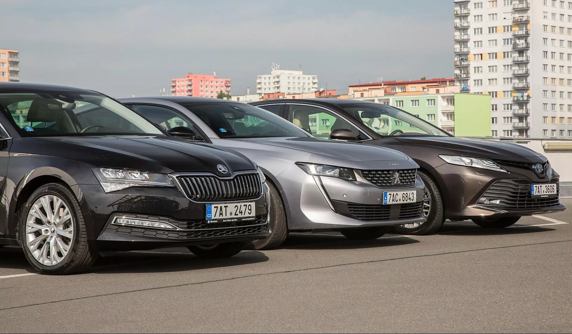 Três carros alinhados lado a lado num estacionamento, fotografados de um ângulo frontal-esquerdo destacando os designs modernos das suas frentes, com um Škoda, apresentando uma grade proeminente e faróis elegantes, um Peugeot com grade distintiva e faróis LED afiados, e um carro parcialmente visível com faróis inclinados para trás, todos mostrando tecnologias de iluminação modernas padrão e designs aerodinâmicos contra um cenário urbano.