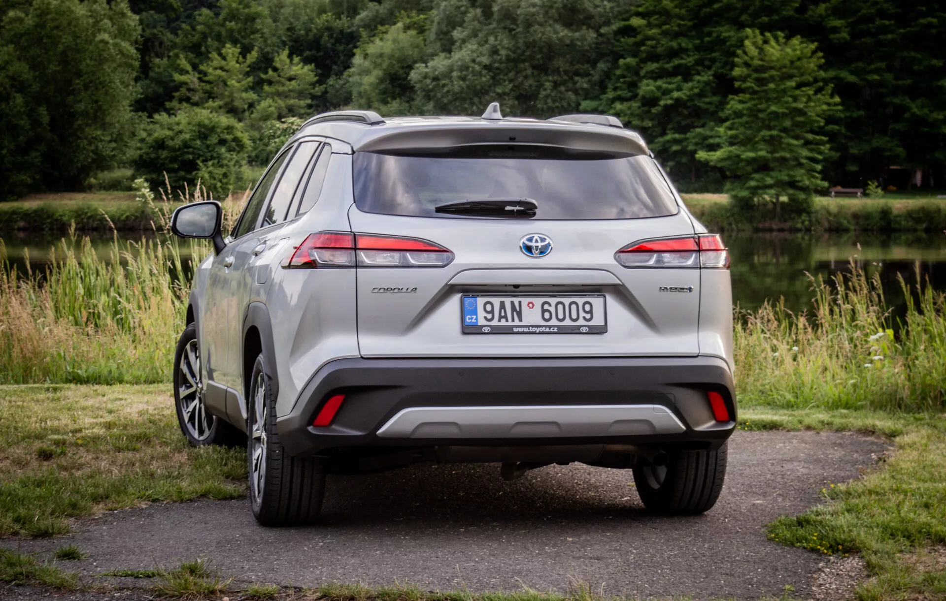 Toyota Corolla Cross prateado de 2022 estacionado em pavimento com vegetação ao fundo exibindo suas luzes traseiras LED, spoiler traseiro, roda de liga com múltiplos raios e emblema híbrido à luz natural do dia