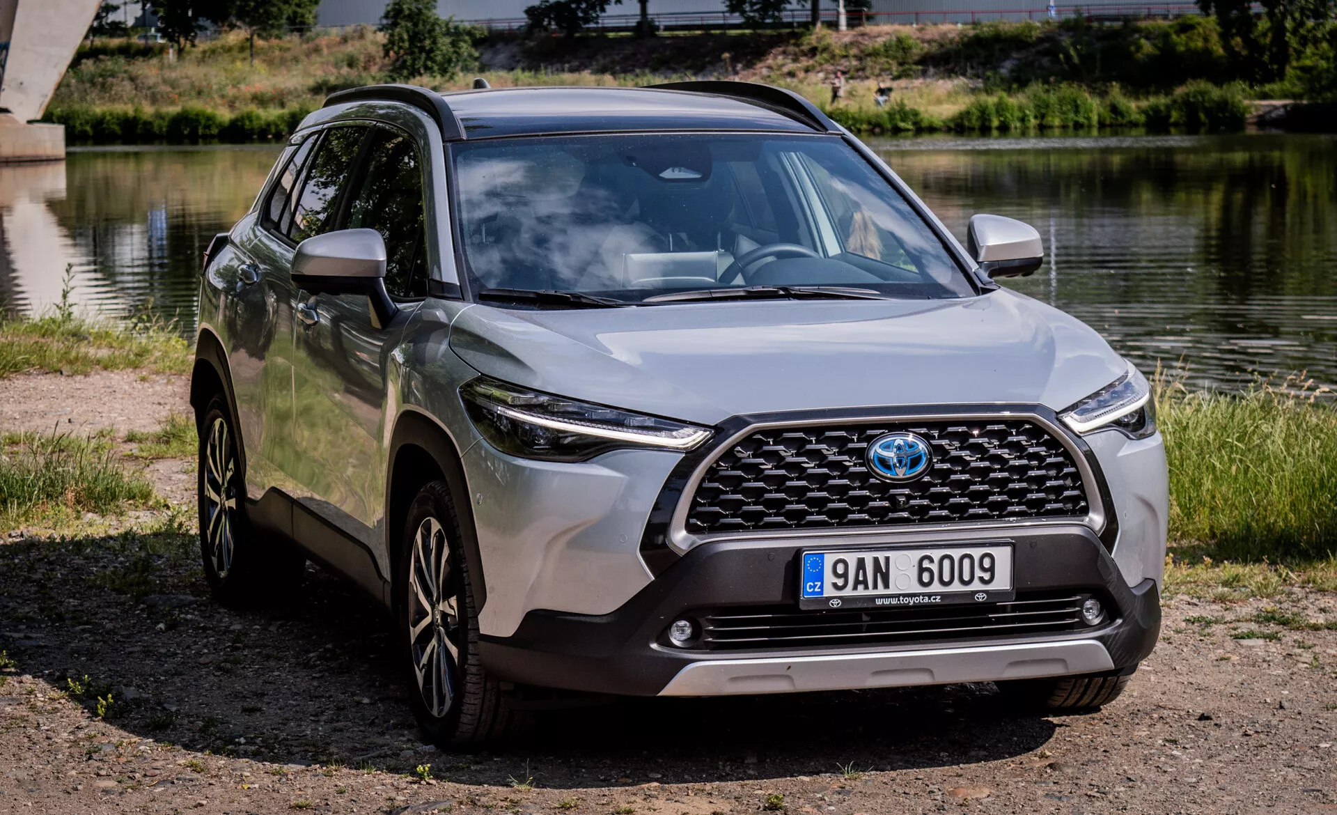 SUV crossover moderno Toyota Corolla Cross 2022 estacionado ao ar livre perto de um corpo de água com vista clara da sua grade frontal, capô, faróis LED, para-choque dianteiro e perfil lateral incluindo rodas de liga leve e retrovisores laterais na cor da carroceria