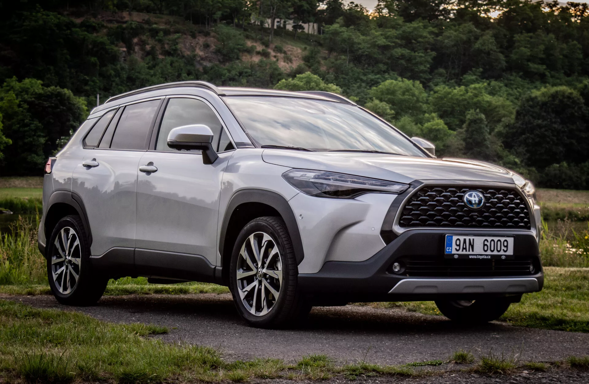 Toyota Corolla Cross 2022 com acabamento metálico brilhante mostrando seu design moderno de SUV com grade frontal detalhada, emblema, faróis LED angulares, para-choque dianteiro aerodinâmico, capô contornado, perfil lateral com rodas de liga leve de múltiplos raios e barras no teto, tirada de um ângulo baixo para enfatizar sua altura e postura durante o dia em um ambiente suburbano natural.