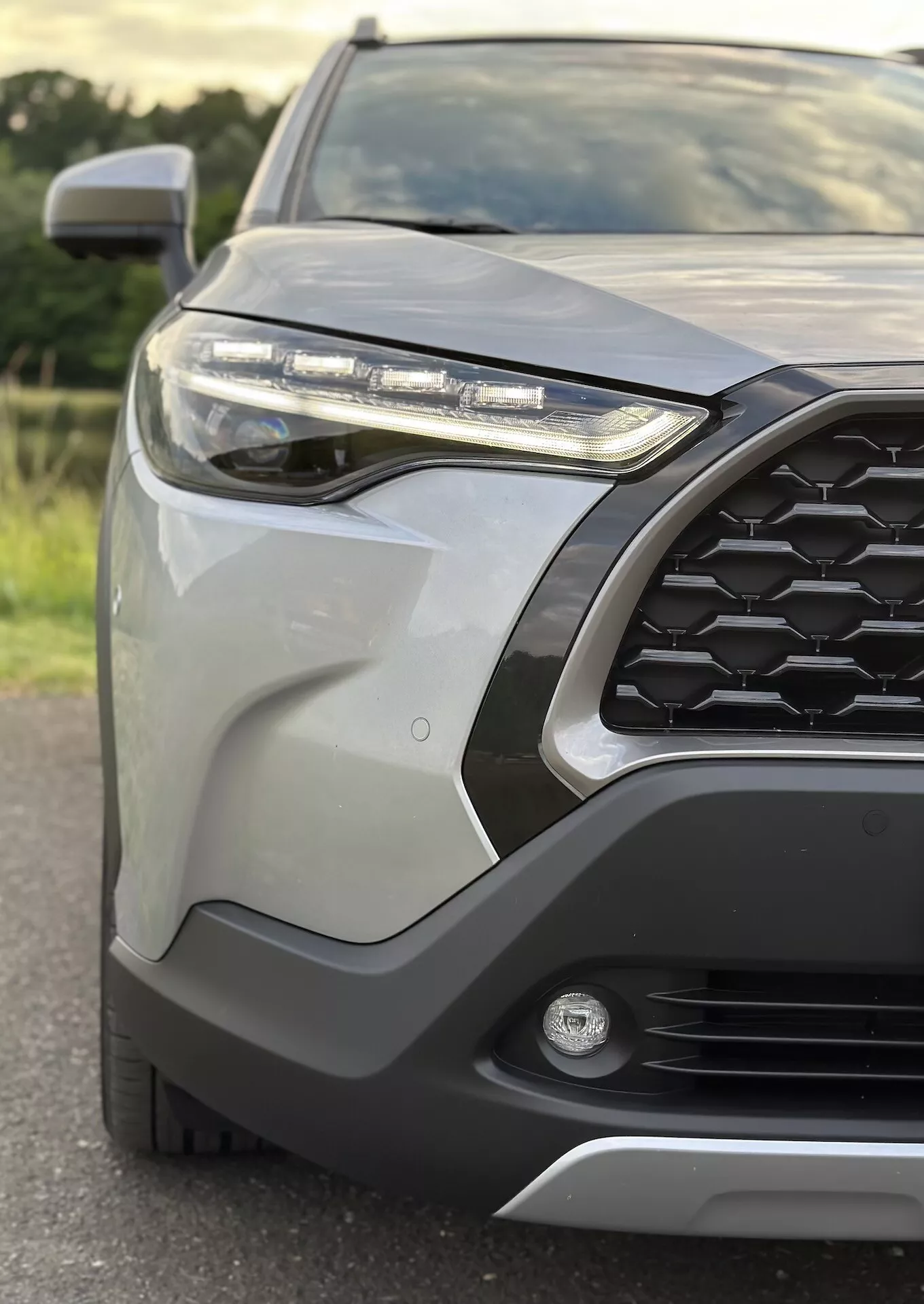 Close-up do quarto frontal-esquerdo do Toyota Corolla Cross 2022 destacando padrão de grade hexagonal, farol LED, luz de neblina, para-choque frontal com revestimento protetor, painéis de carroceria elegantes, espelho lateral com sinalizador integrado e acabamento metálico
