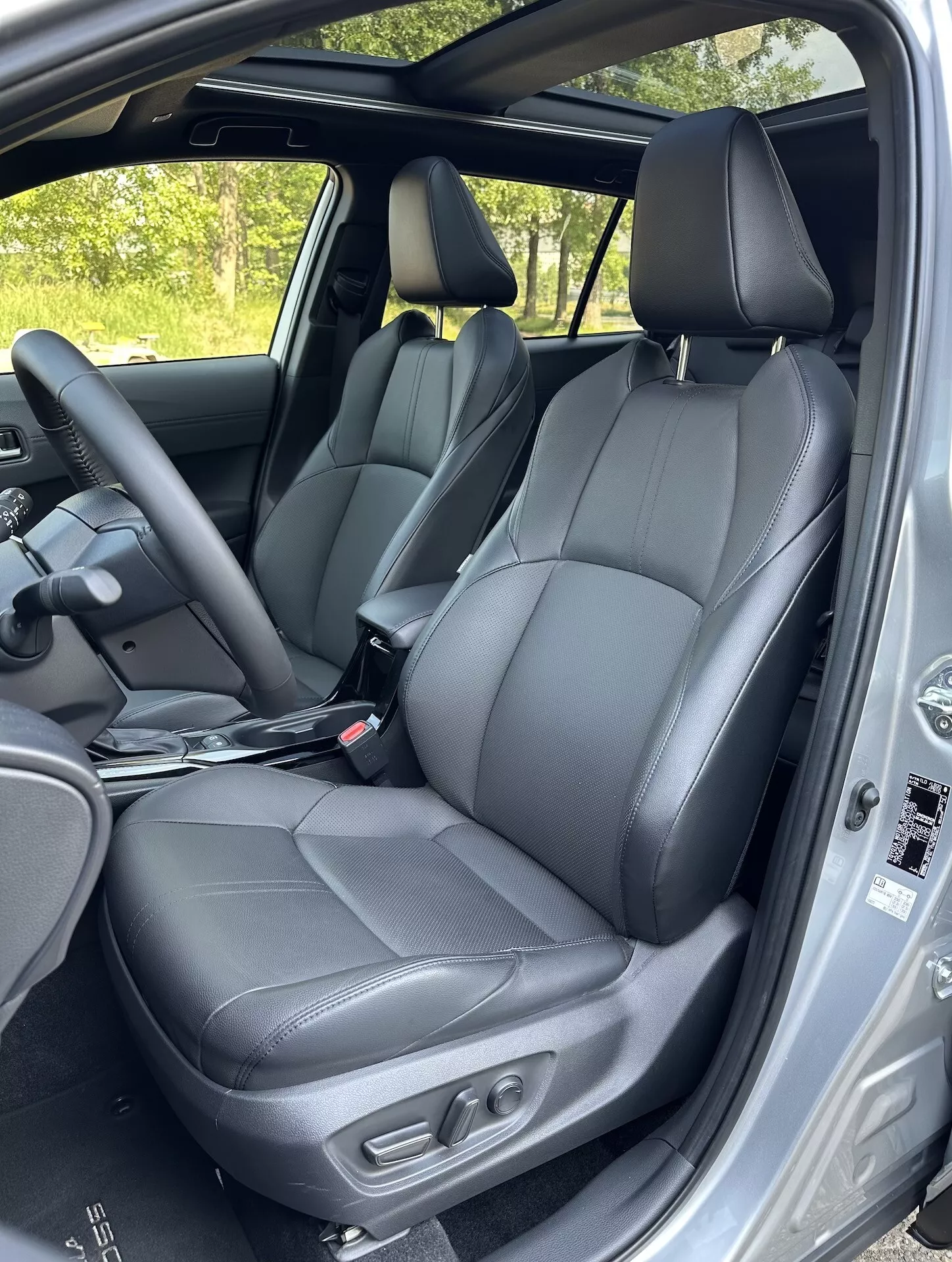 Vista interior do Toyota Corolla Cross 2022 destacando os assentos dianteiros em couro preto com controles de ajuste, parte dos assentos traseiros, volante e características de segurança da moldura da porta à luz natural do dia