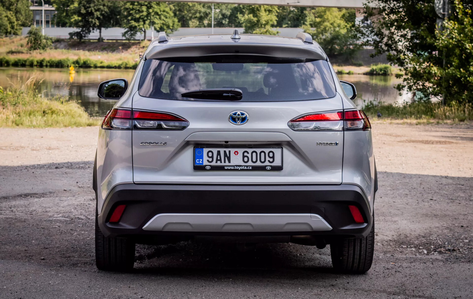 Vista traseira do Toyota Corolla Cross 2022, exibindo lanternas traseiras LED, emblema híbrido e emblema cromado da Toyota com iluminação natural diurna e um cenário pitoresco de vegetação e água
