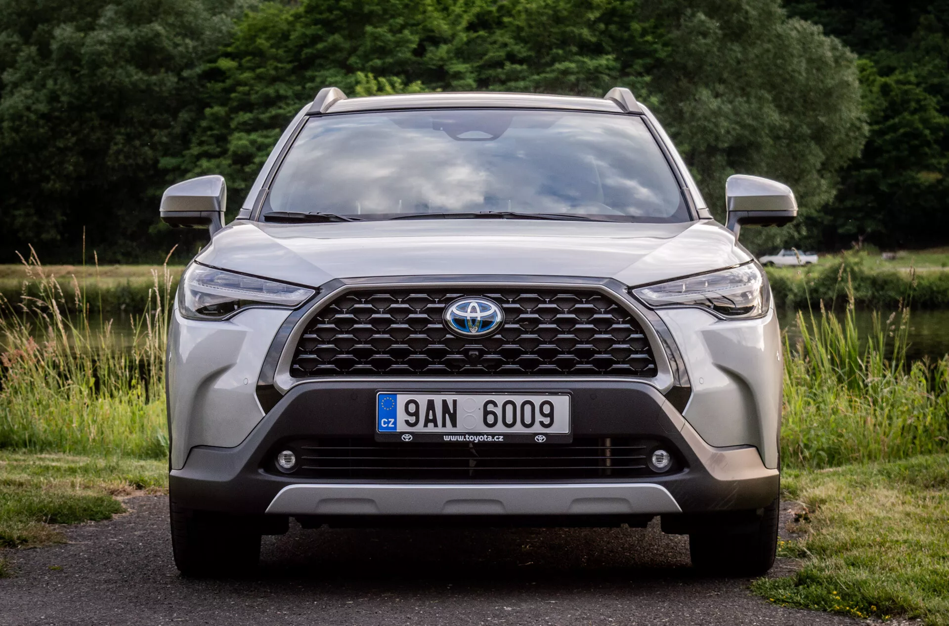 Vista frontal de um Toyota Corolla Cross 2022, destacando sua grade grande com emblema, faróis modernos de LED, luzes de nevoeiro, posição da placa de licença, sensores de estacionamento integrados ou câmera frontal, contornos aerodinâmicos do capô, design robusto do para-choque inferior e entradas de ar, estacionado no asfalto com um fundo natural