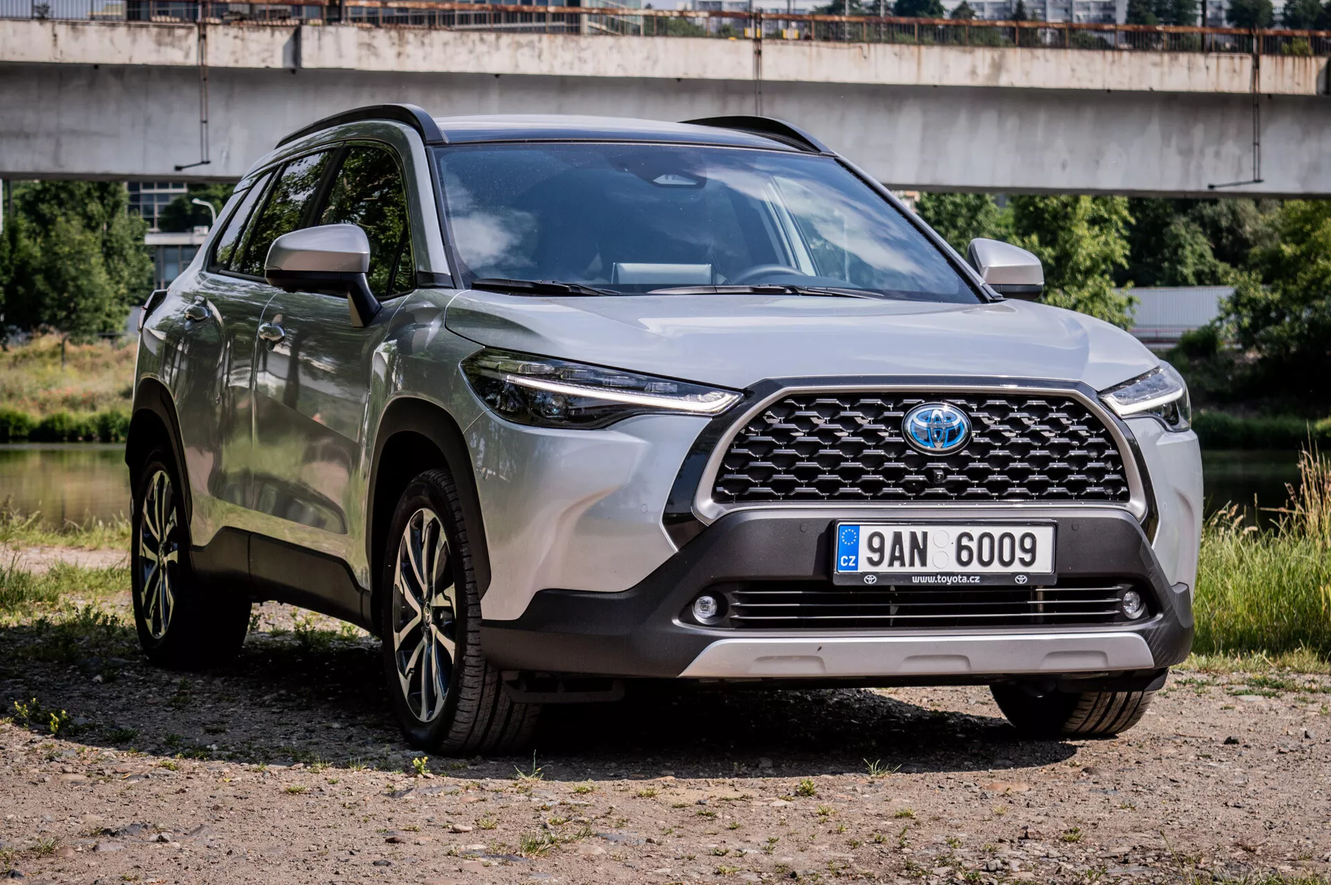 Toyota Corolla Cross 2022 estacionado em superfície de cascalho exibindo seu design robusto, rodas de liga leve esportivas de múltiplos raios, faróis de LED avançados e alta distância do solo em vista frontal de três quartos destacando a grade frontal, para-choque e características do lado do motorista com uma ponte ao fundo em ambiente semiurbano