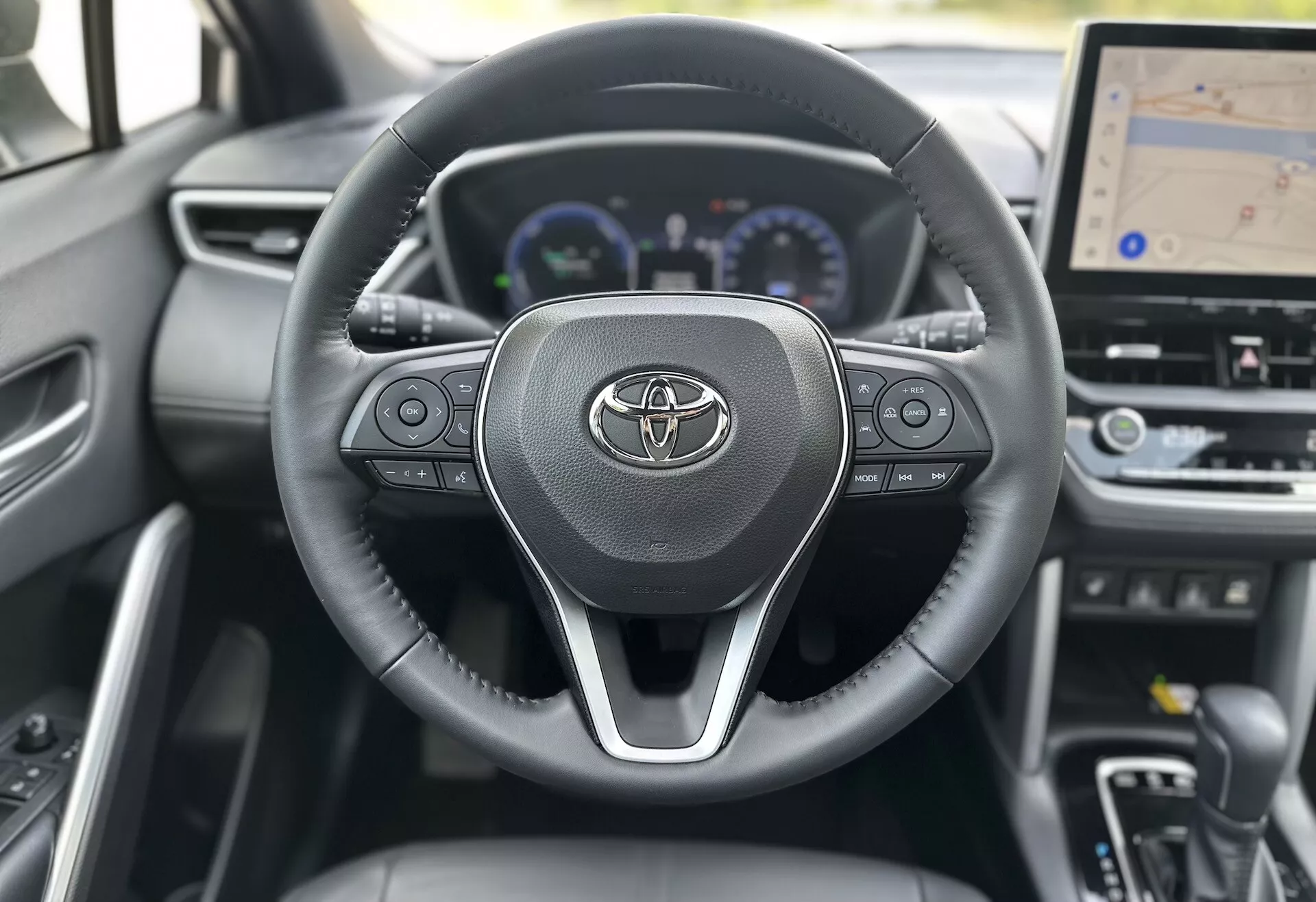 Interior do Toyota Corolla Cross 2022, mostrando o volante com o logotipo da Toyota, controles integrados para áudio, chamadas e controle de cruzeiro, atrás dos quais um painel digital com velocímetro, tacômetro e display multifuncional é parcialmente visível, incluindo um display central maior para navegação GPS, tudo em um fundo de design contemporâneo em materiais escuros.
