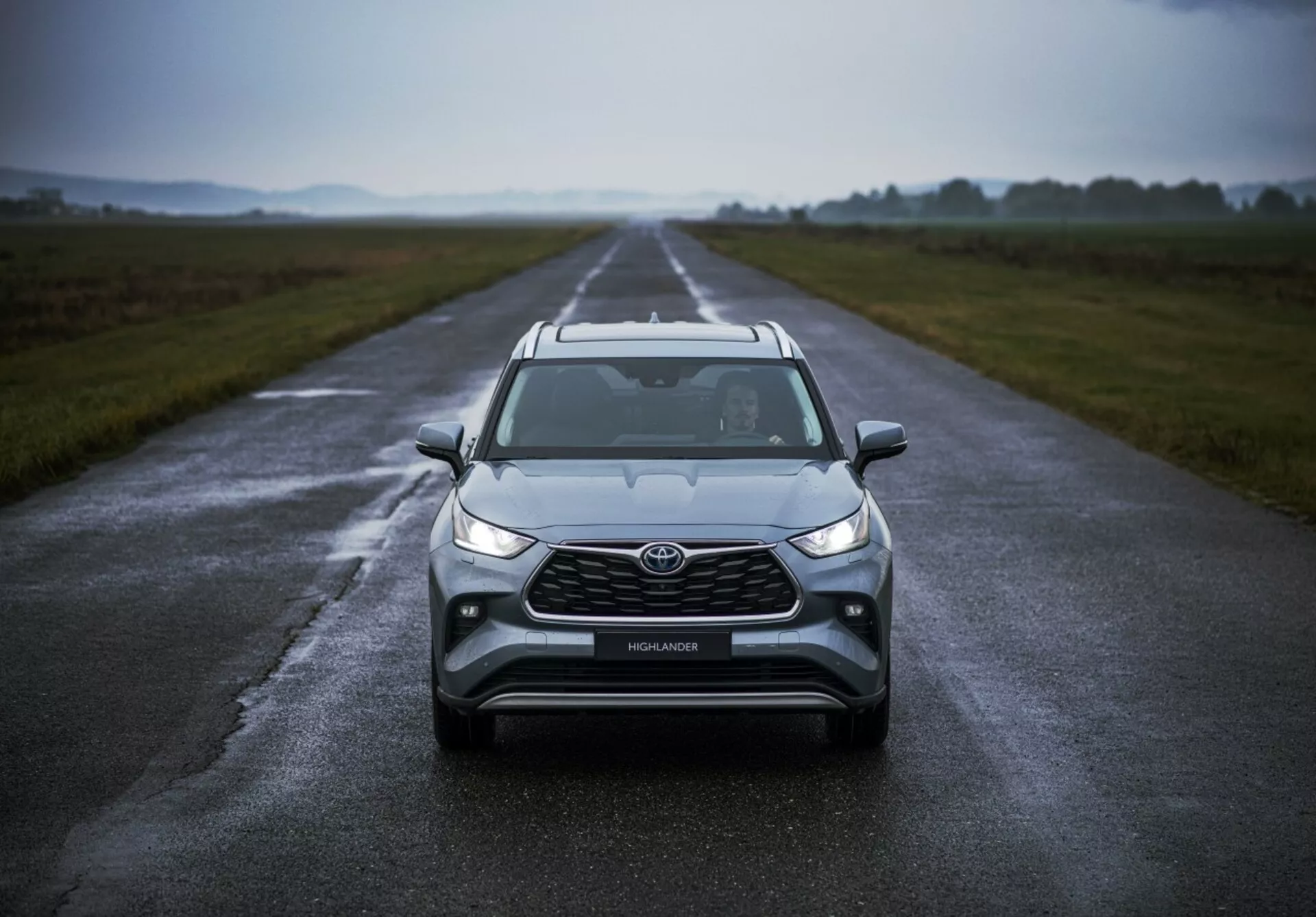 Toyota Highlander 2021 posicionado em uma estrada molhada destacando seu design frontal, clima nublado, com faróis de LED, luzes de neblina, uma grade grande e tecnologia de segurança avançada