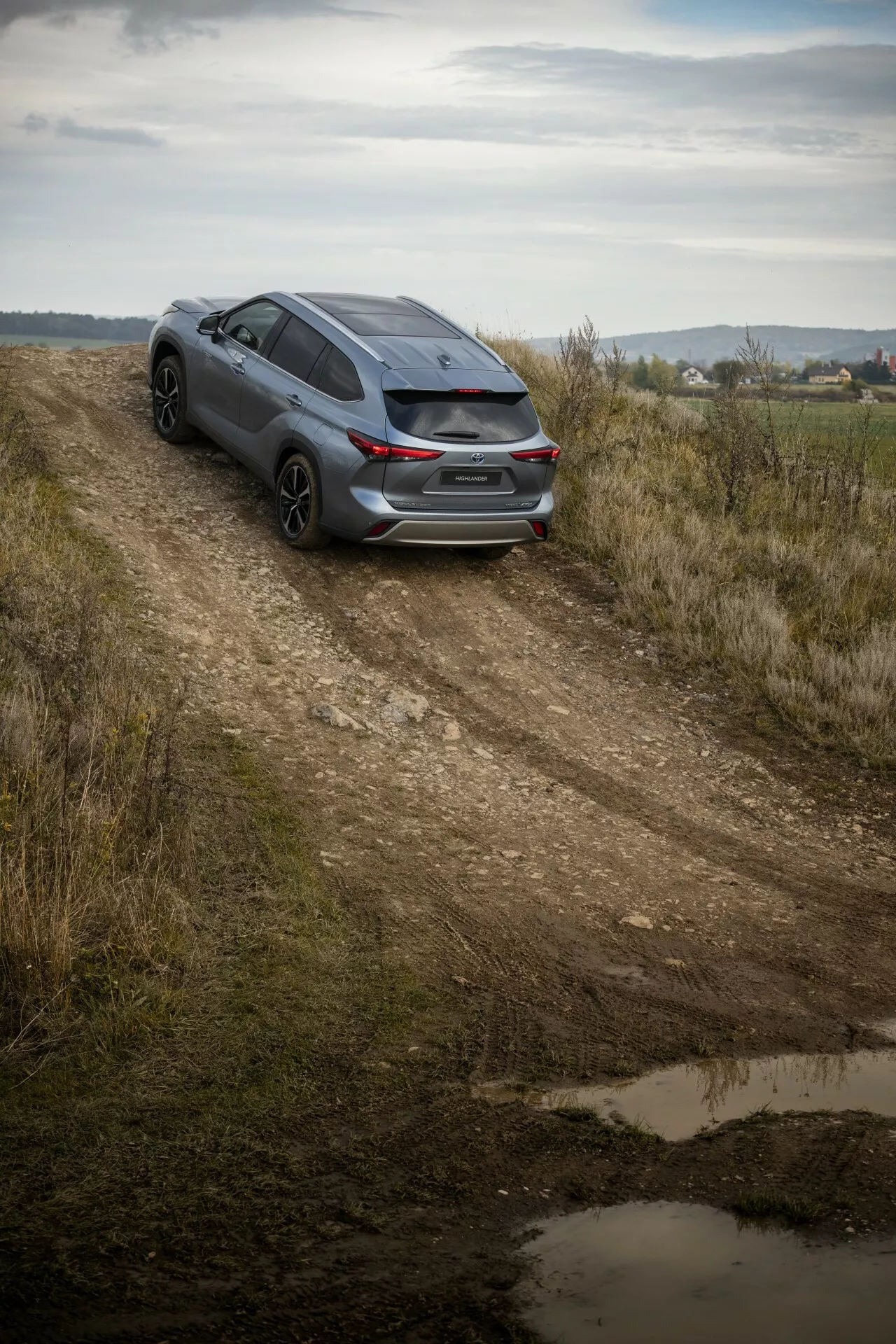 Toyota Highlander 2021 escuro capturado de um ângulo alto em uma estrada de terra, exibindo a vista traseira de três quartos, design esportivo e potencial para viagens leves em todo terreno em meio a uma paisagem campestre