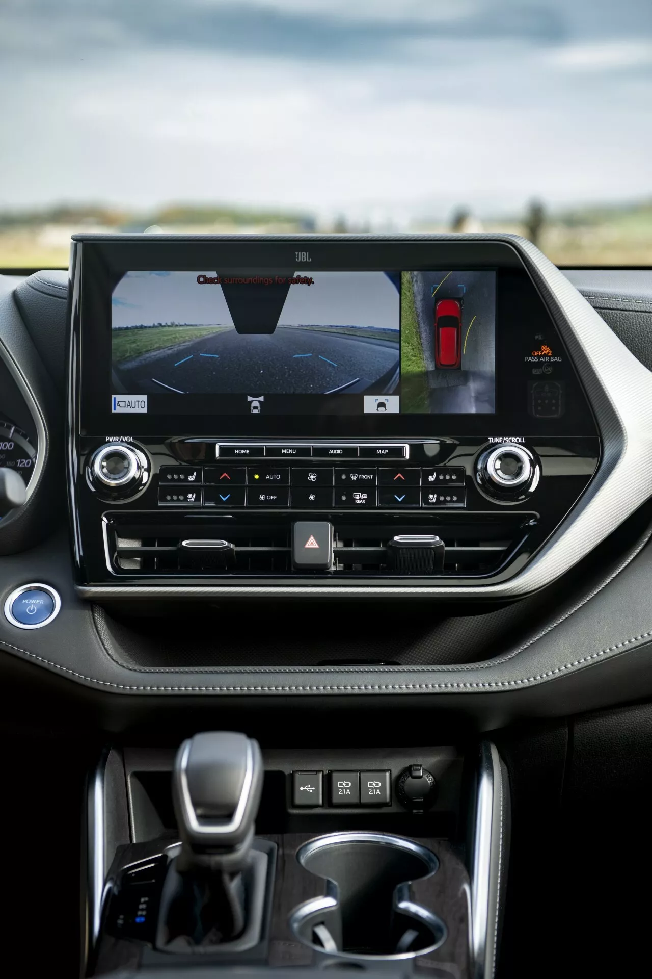 Interior do Toyota Highlander 2021 destacando a consola central com tela sensível ao toque orientada para a paisagem exibindo câmera traseira e assistente de estacionamento, controles de clima de zona dupla e alavanca de transmissão automática