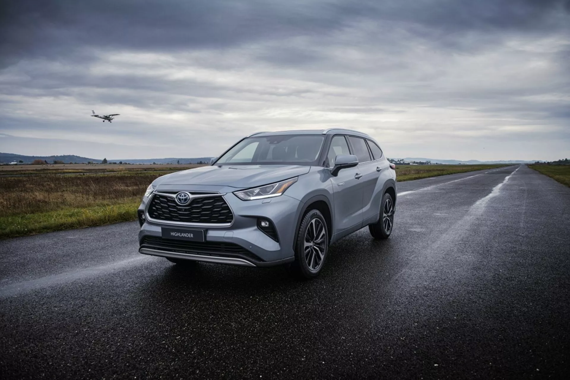 SUV crossover Toyota Highlander 2021 em estrada de asfalto molhada, exibindo seus faróis angulares, grade frontal proeminente, rodas de liga leve multirradiais e design aerodinâmico sob iluminação difusa e nublada