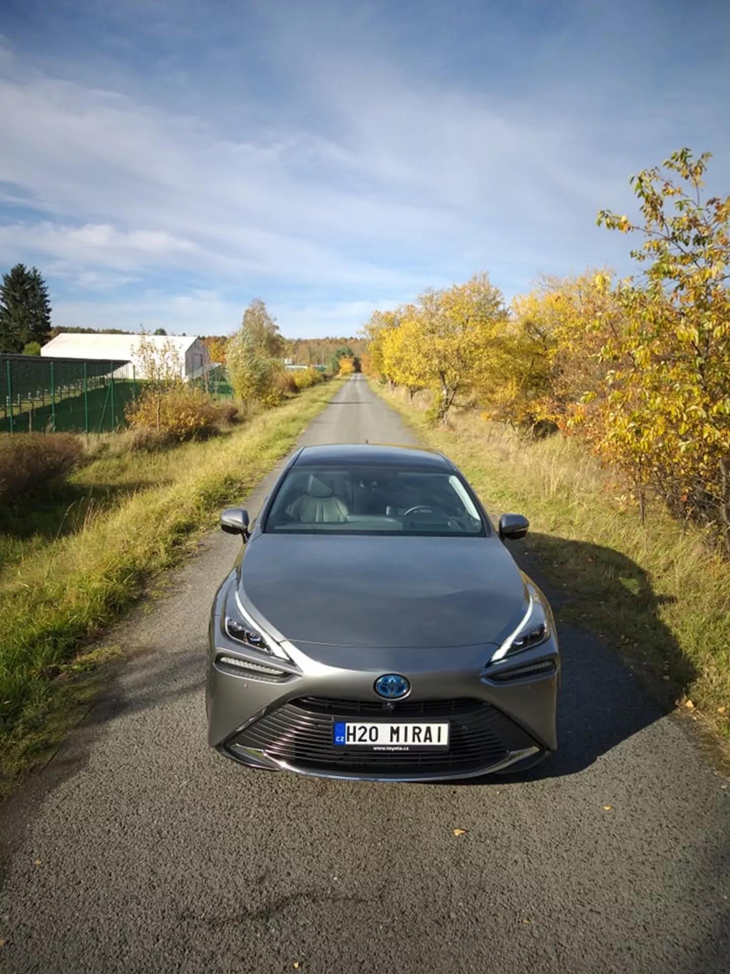 Toyota Mirai 2021 fotografado de uma perspectiva frontal e alto ângulo, destacando seus faróis LED elegantes e grade proeminente com emblema central que pode incluir tecnologia de sensores, em um contexto semi-rural sob luz natural, mostrando a avançada tecnologia de célula de combustível de hidrogênio e design aerodinâmico
