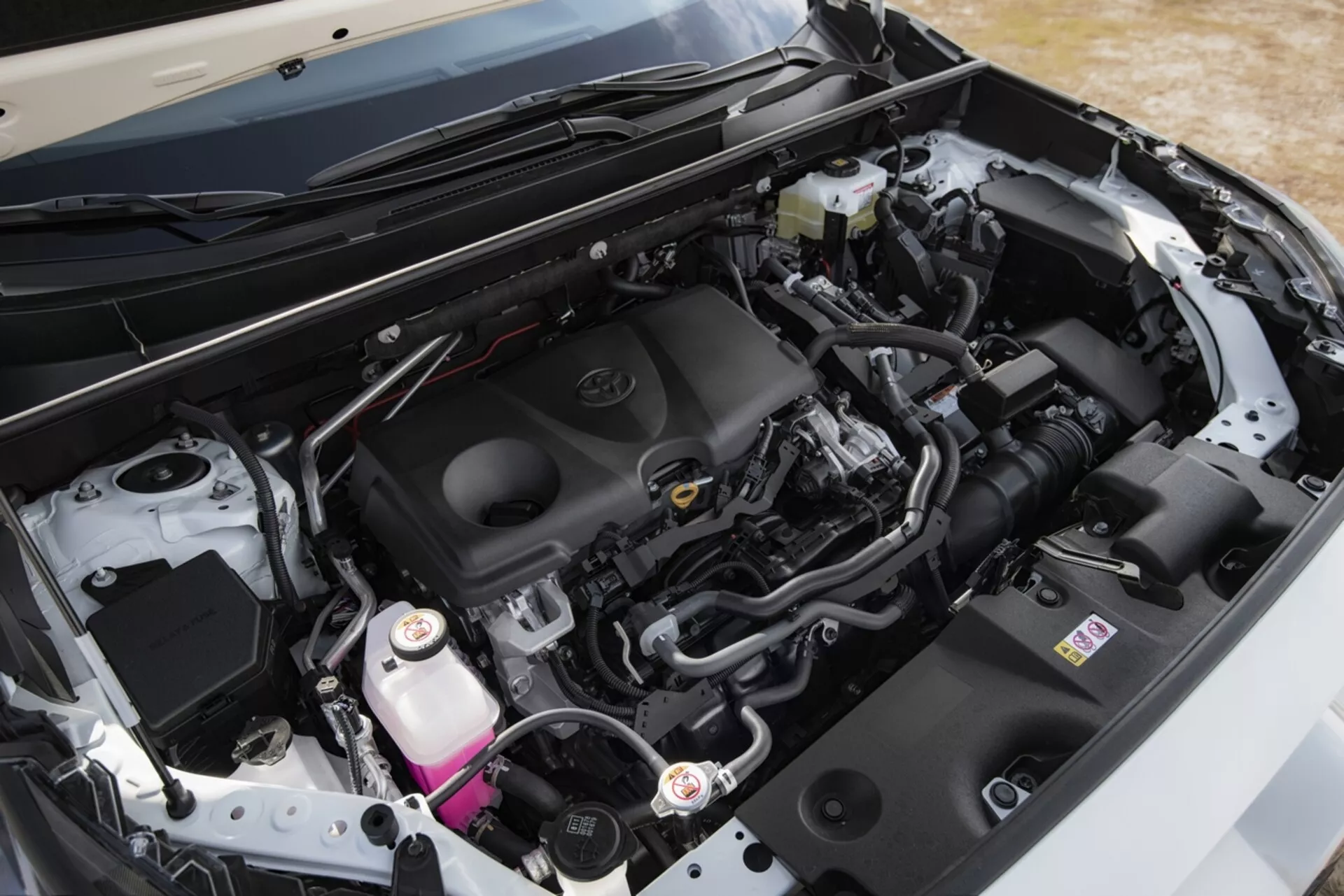 Vista de perto do compartimento do motor de um Toyota RAV4 2019, destacando o design limpo e moderno e componentes como a tampa do motor com emblema da marca, sistema de admissão de ar, bateria, reservatório de fluido de freio, e reservatório de refrigerante, ao lado de vários tubos e chicotes de fios, sob o capô aberto de um ângulo oblíquo.