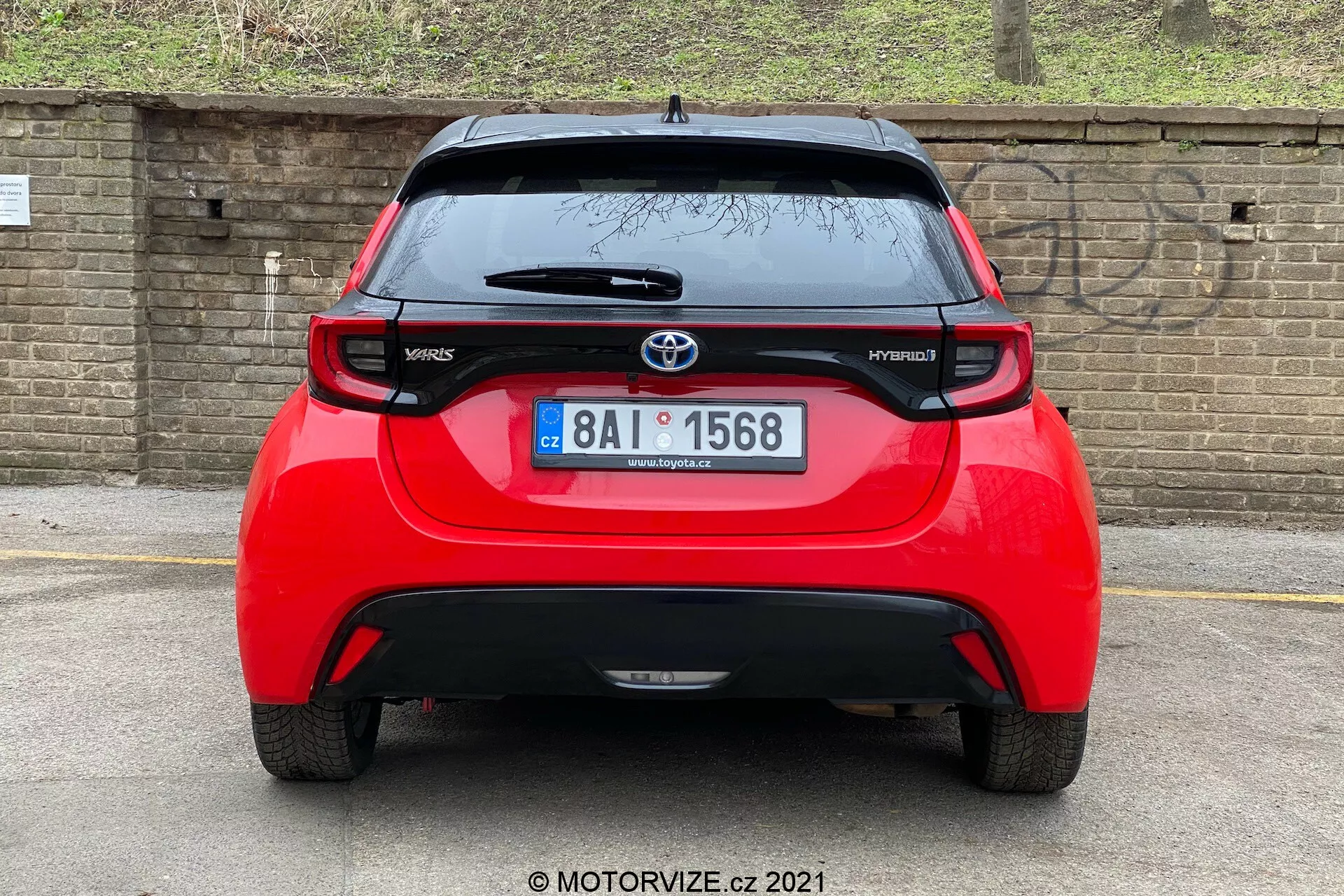 Vista traseira de um Toyota Yaris Hybrid vermelho de 2020, centrado e tirado em um ângulo direto. A imagem revela toda a largura do veículo, mostrando o emblema da Toyota, os nomes dos modelos 'Yaris' e 'Hybrid', um spoiler, antena montada no teto, faixa de luz de freio alta e luzes traseiras distintas. O vidro traseiro com limpador e a parte inferior do pára-choques, que pode abrigar sensores ou refletores, também são visíveis. O fundo inclui uma parede de tijolos ou cerca e alguma vegetação, sugerindo um carro estacionado mais baixo ou um ângulo de câmera mais baixo.