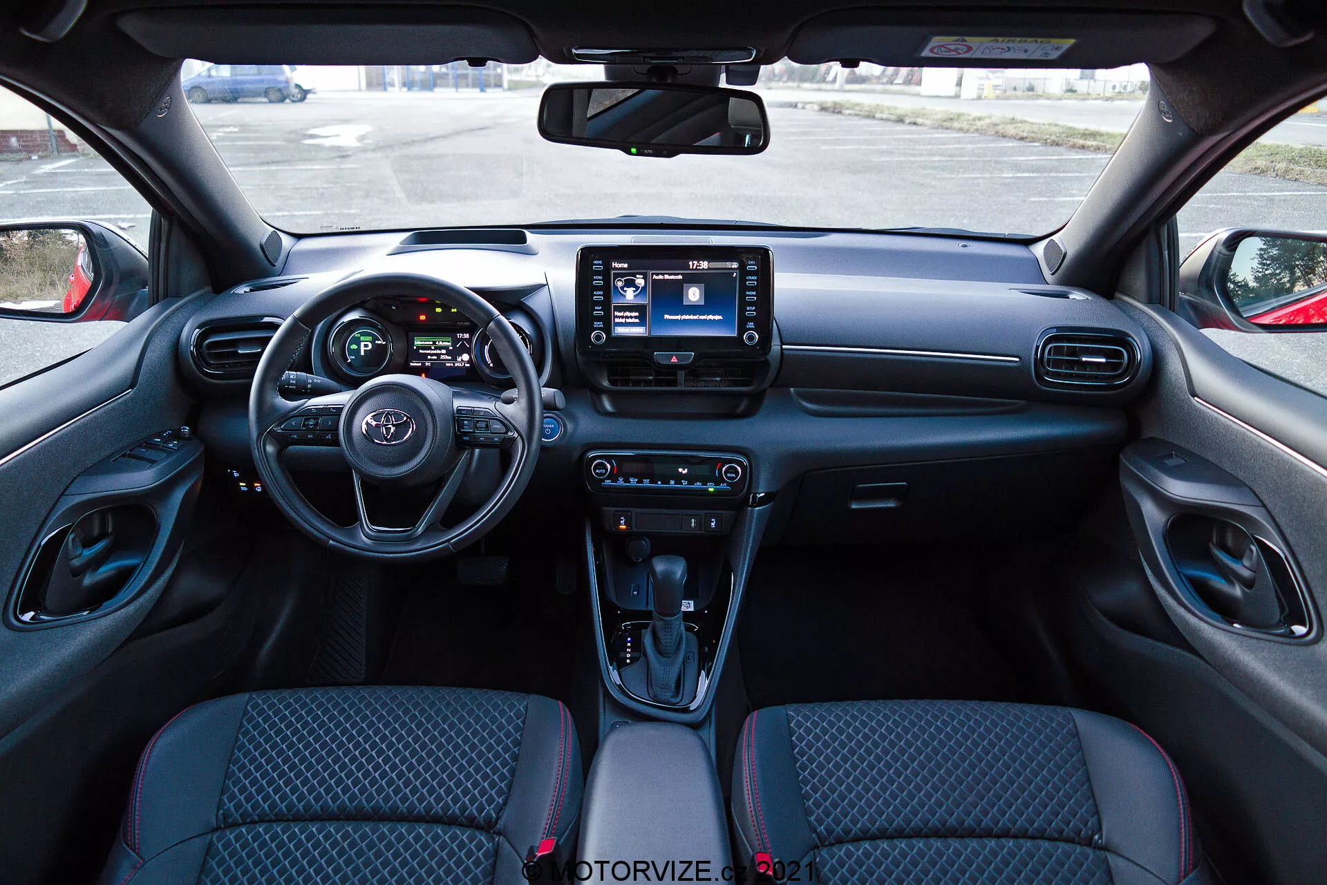 Vista interior de um Toyota Yaris 2020 a partir da perspectiva do assento do motorista, com um painel moderno que apresenta um volante com controles integrados, um painel de instrumentos parcialmente visível com telas digitais e um sistema de infotainment central com interface touchscreen. O painel inclui saídas de ar e botões de controle de clima, enquanto o console central possui controles para modos de condução, uma alavanca de câmbio automática com capa tipo couro, porta-copos e um compartimento de armazenamento. Os assentos estofados em tecido têm um design estampado e os painéis interiores das portas incluem controles para janelas elétricas e ajustes dos espelhos.