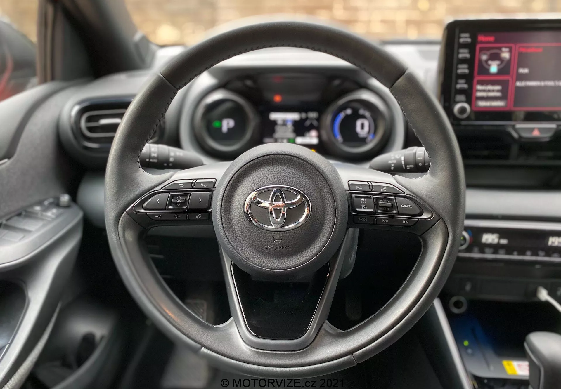 Close-up do interior do Toyota Yaris 2020 visto do assento do condutor, com volante de três raios com emblema da Toyota e botões de controle para áudio, chamadas em modo mãos-livres e controle de cruzeiro. O painel de instrumentos digital que mostra velocidade e informações essenciais de condução é parcialmente visível. O console central com tela sensível ao toque do sistema de infoentretenimento, exibindo opções de menu, também é visível.