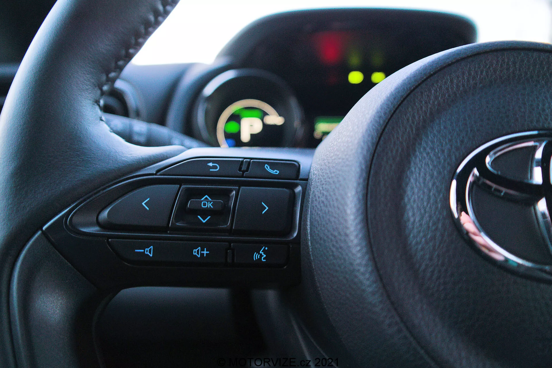 Vista interior aproximada de um Toyota Yaris 2020 a partir da perspectiva do assento do motorista, destacando o volante com controles multifuncionais e um painel de instrumentos ao fundo. O volante domina os dois terços inferiores da imagem, com setas, comandos de voz, controles de volume e botões do sistema de entretenimento visíveis. O painel de instrumentos parcialmente visível está iluminado com um display digital verde e está fora de foco devido à baixa profundidade de campo. A luz natural ilumina o interior, enfatizando o conforto e uma interface amigável.