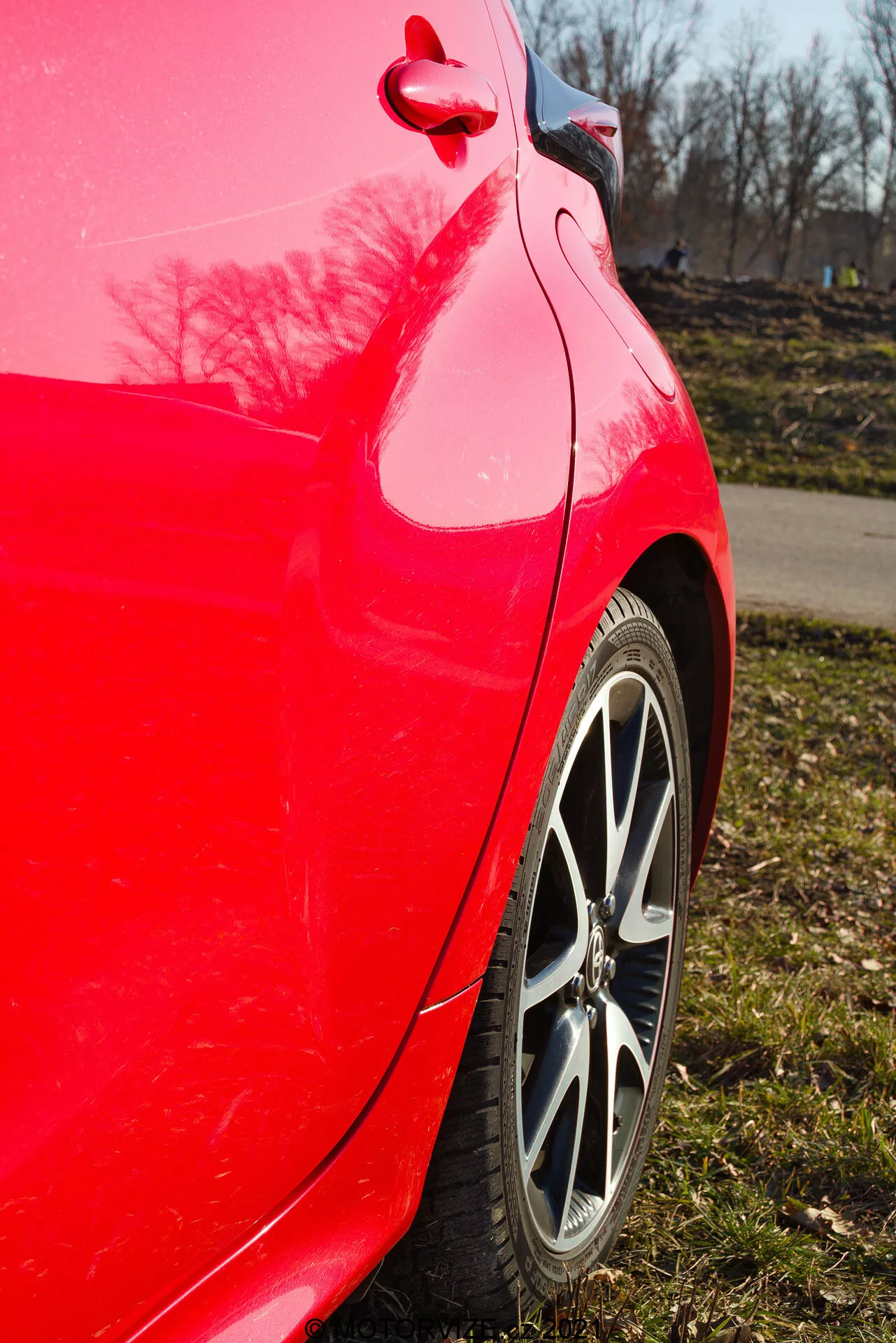 Vista lateral de perto do painel traseiro de um Toyota Yaris 2020 vermelho, tirada em um ângulo ligeiramente inclinado para cima, destacando o acabamento vermelho brilhante, a jante de liga de múltiplos raios, o padrão do piso do pneu, a curvatura limpa do painel traseiro e do para-choque, o reflexo brilhante da pintura com árvores e um céu azul claro, e a tampa de combustível e detalhe de acabamento visíveis.