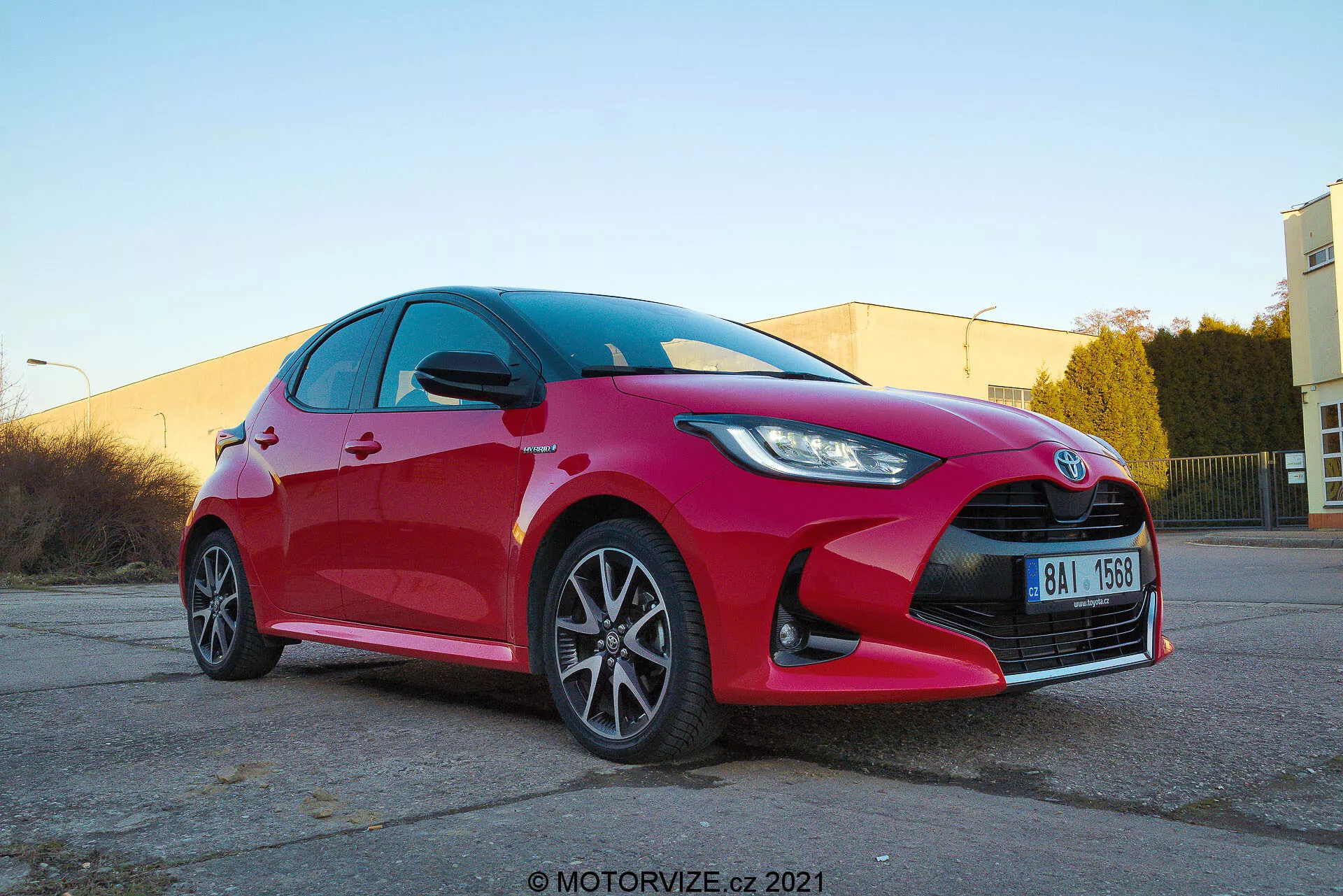 Vista frontal de baixo ângulo de três quartos de um vibrante Toyota Yaris 2020 hatchback vermelho, mostrando faróis LED ou HID angulares e nítidos, grade proeminente com emblema central, rodas de liga leve de múltiplos raios, indicadores de direção integrados nos espelhos laterais, superfícies refletivas limpas e um possível sistema de freios orientado para desempenho. Capturado em uma área de estacionamento industrial iluminada por uma luz quente e com marca d'água motorvize.cz 2021.