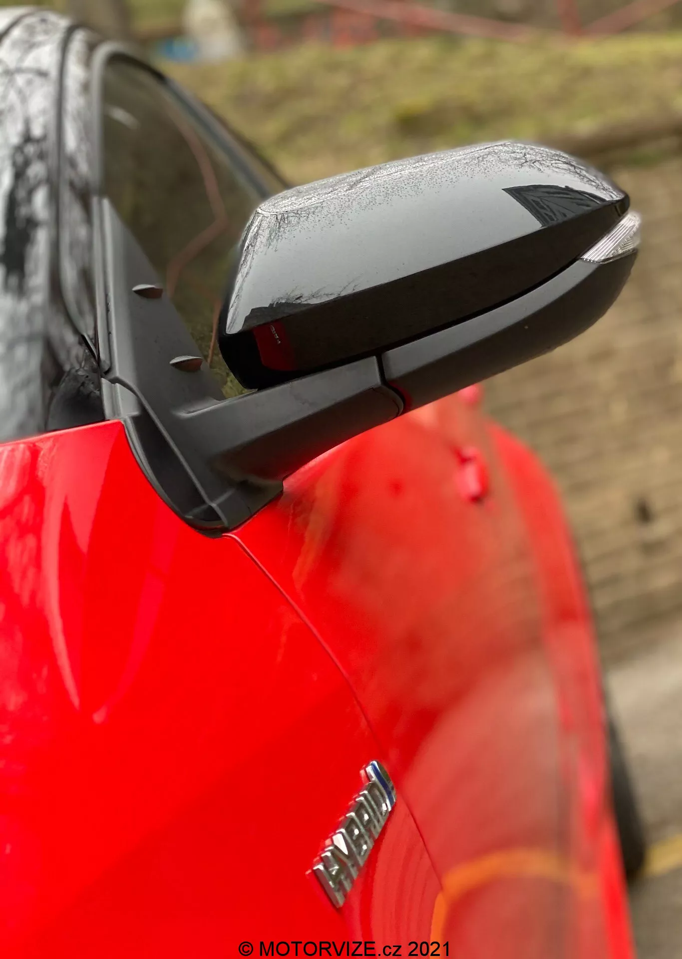 Close-up do espelho lateral desdobrado de um Toyota Yaris 2020, com uma carcaça preta aerodinâmica elegante que contrasta com a carroceria vermelha brilhante do carro. O espelho destaca toques tecnológicos possíveis como ajuste elétrico, aquecimento e sinais de direção integrados, embora características específicas não sejam confirmadas. Gotas de água sugerem chuva recente ou lavagem do carro, enquanto um fundo desfocado e uma baixa profundidade de campo mantêm o foco no espelho. Parte de um emblema no para-lama dianteiro é visível, mas desfocado.