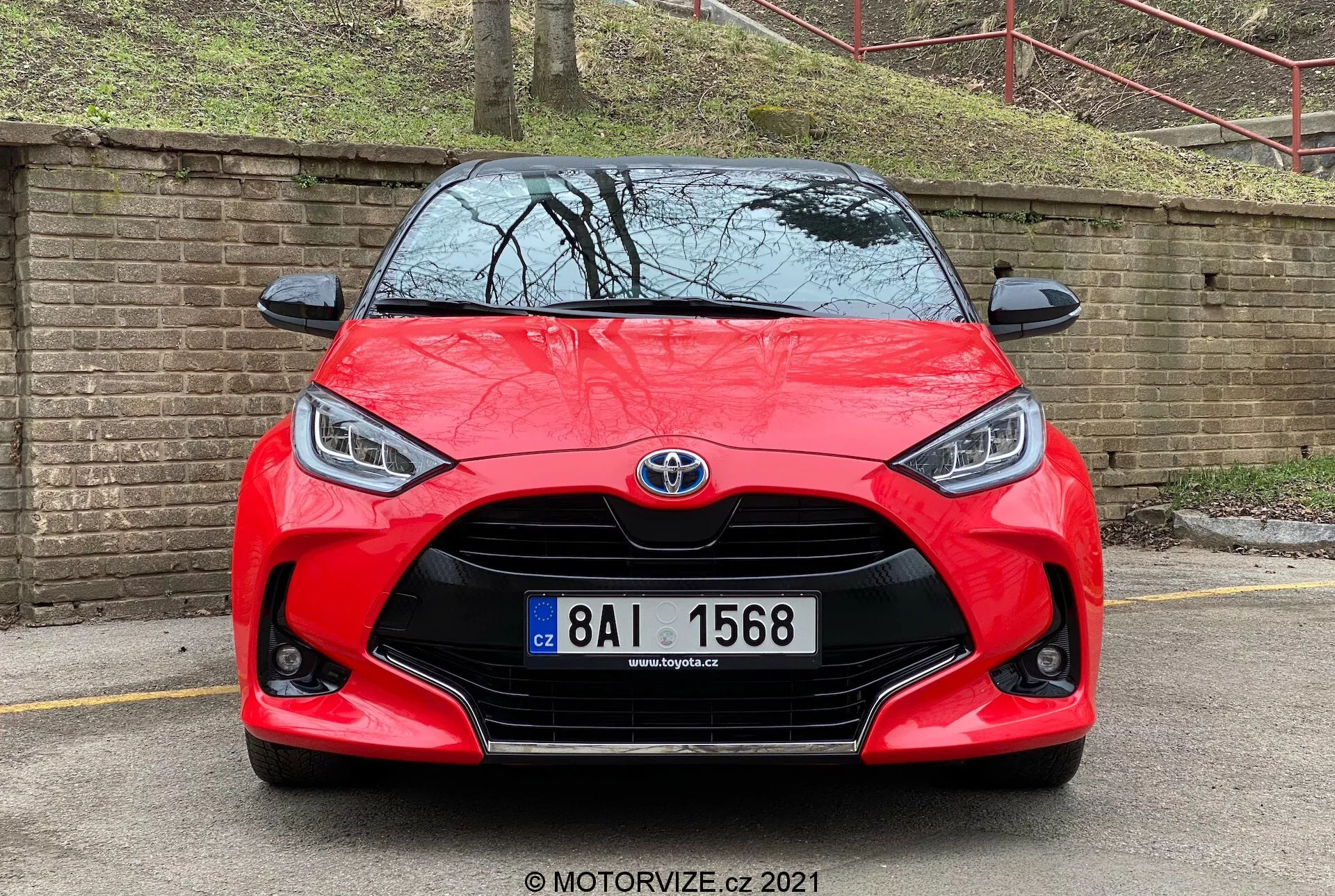 Vista frontal de um Toyota Yaris vermelho 2020, exibindo sua distinta grade frontal com um emblema Toyota proeminente no centro. A grade em estilo de malha é ladeada por faróis angulares elegantes que apresentam luzes diurnas de LED. Abaixo da grade, há uma entrada de ar visível cercada por casas de faróis de neblina. O capô esculpido e o para-choque dianteiro perfeitamente integrado contribuem para o design esportivo do carro. A placa de licença está montada no centro, e a imagem possui uma marca d'água MOTORVIZE.cz, datada de 2021.
