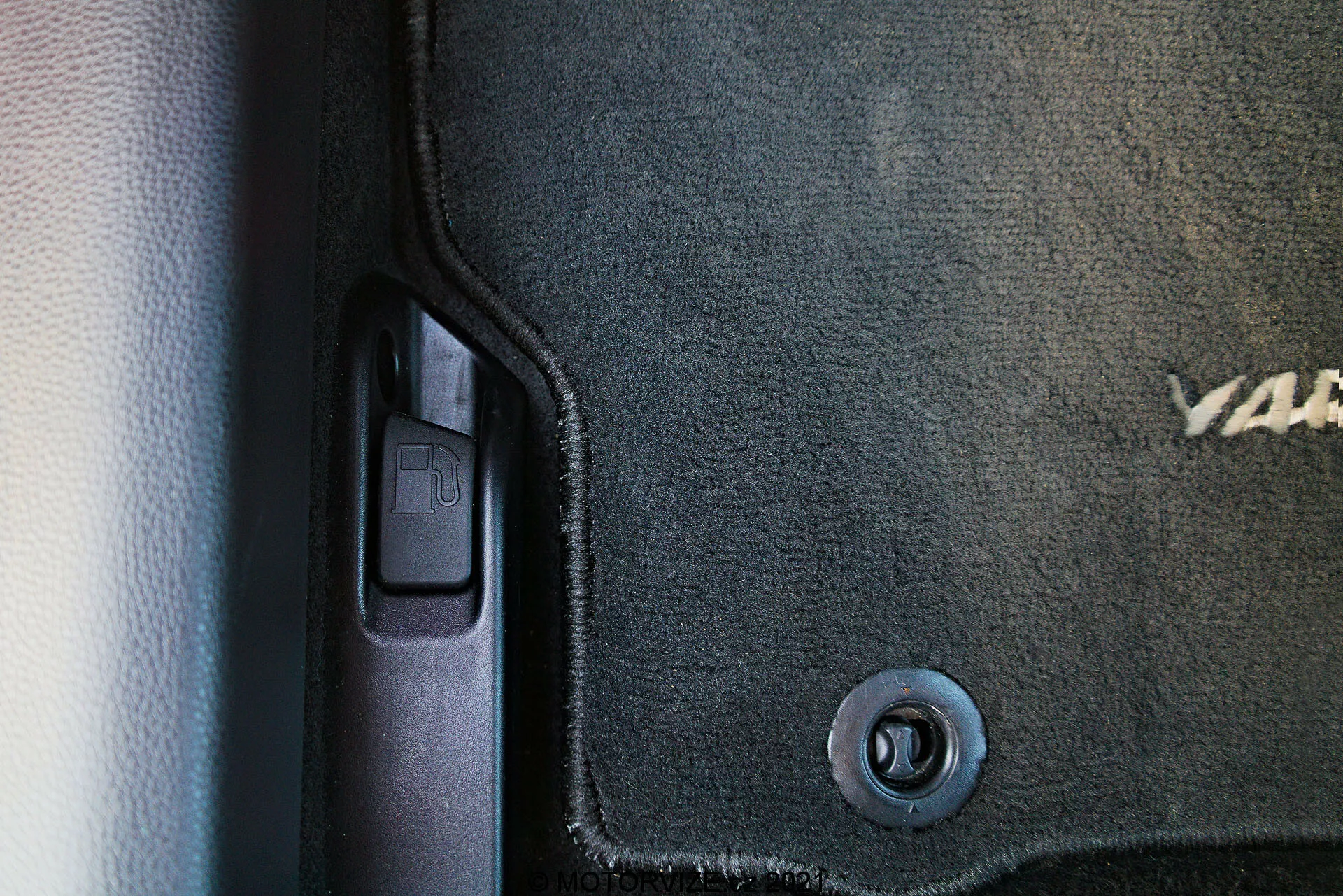 Close-up do interior do Toyota Yaris 2020, capturando o lado inferior esquerdo próximo à porta. Visíveis estão um tapete de cor escura com texto bordado parcial, um gancho de retenção circular de metal, o batente de porta de plástico com uma moldura protetora, uma alavanca de abertura do porta-malas sugerindo o lado do motorista e um esquema de cores em dois tons com texturas contrastantes de tapete e plástico liso.