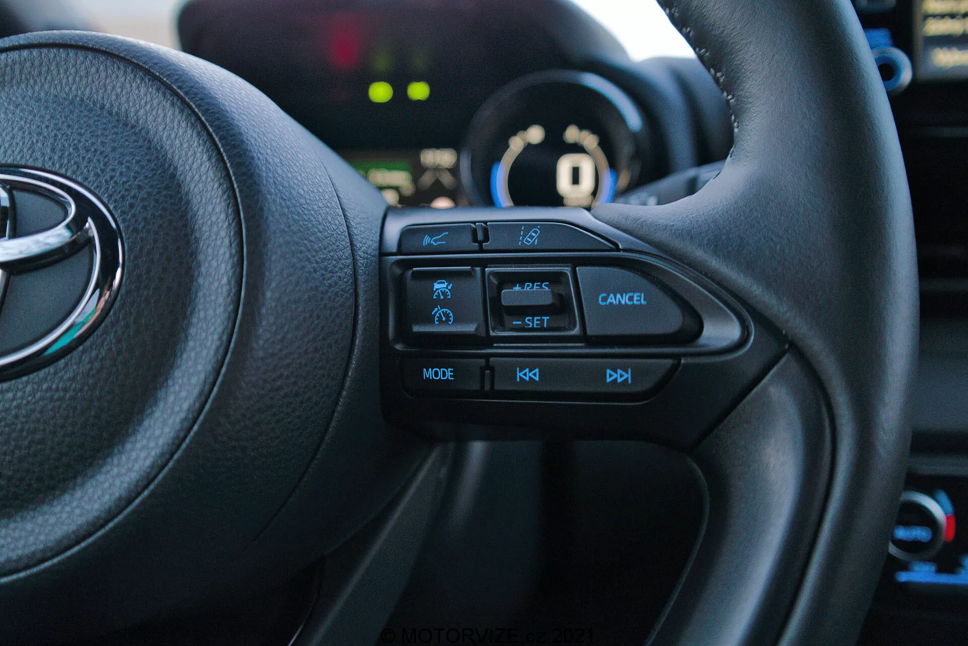 Imagem do interior de um Toyota Yaris 2020 a partir do banco do condutor, focando-se no lado esquerdo do volante multifuncional. Os botões táteis, incluindo o ícone de recurso de segurança, controle de cruzeiro com etiqueta 'SET', botão de cancelamento, 'MODE' para multimídia e controles de volume '+', '-', estão claramente visíveis. O veículo está em modo 'P' estacionamento, com o painel de instrumentos e elementos de fundo levemente desfocados.