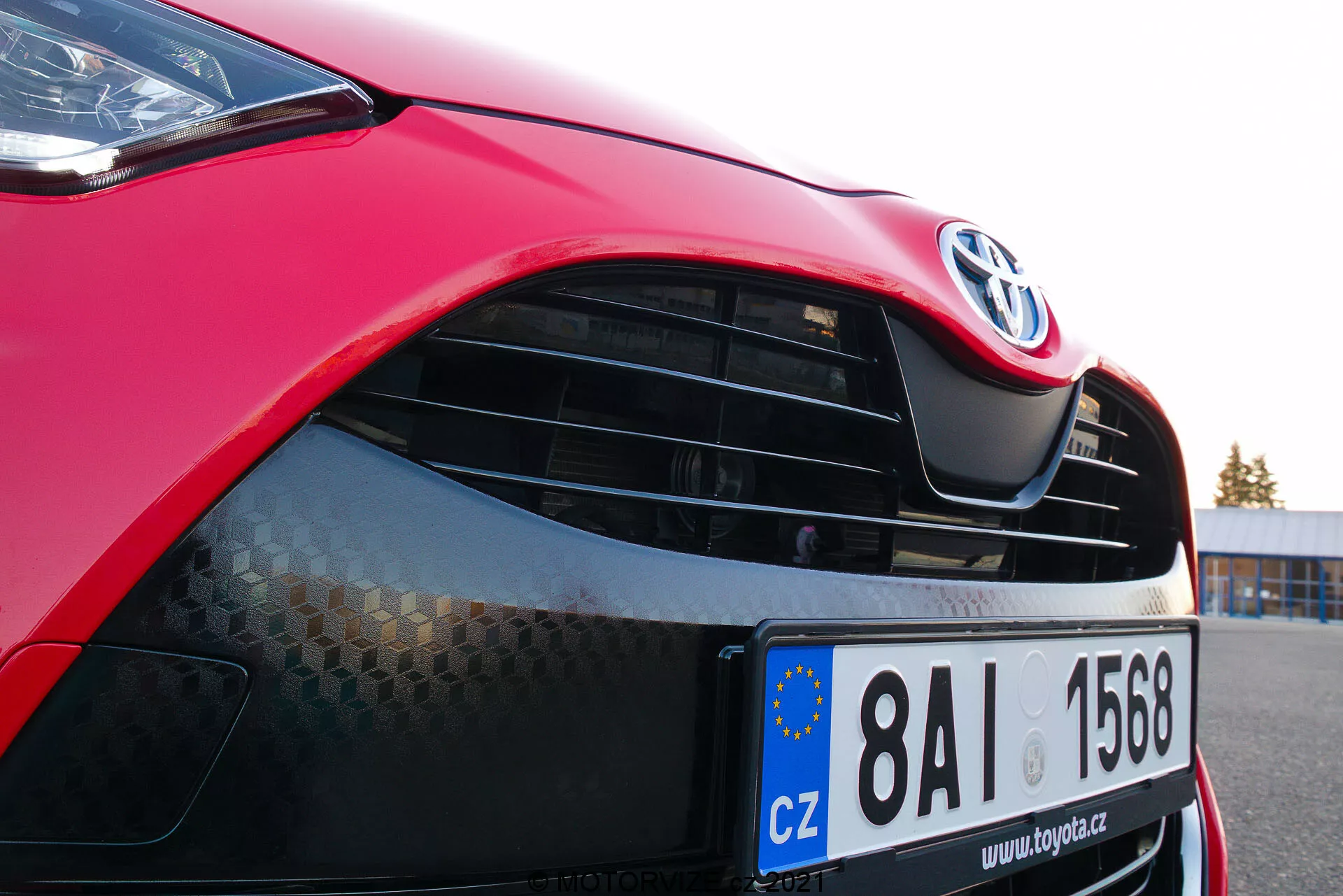 Imagem em close-up de um Toyota Yaris 2020 vermelho enfocando o lado dianteiro esquerdo, mostrando o capô, farol moderno com tecnologia potencial LED ou HID, grade com emblema Toyota e padrão de favo de mel, pintura vermelha brilhante e para-choque dianteiro parcialmente visível com entrada de ar inferior. A placa da União Europeia indica registro na República Checa.
