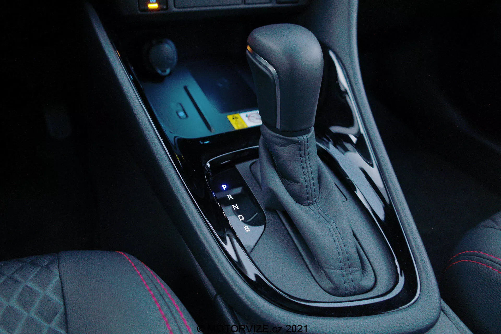 Close-up do seletor de câmbio automático do Toyota Yaris 2020, visto do lado do passageiro frontal em um ângulo ligeiramente inclinado para baixo, destacando o acabamento metálico elegante e o design ergonômico. O seletor de câmbio está posicionado centralmente entre os bancos do motorista e do passageiro frontal e inclui as posições Park, Reverse, Neutral e Drive, bem como opções de mudança manual (+ e -). À frente do seletor estão os controles eletrônicos para modos de condução e sistemas de controle de tração, junto com um sistema de partida por botão iluminado em azul. O console central escuro e brilhante adiciona um toque de luxo moderno, complementado por controles de aquecimento dos bancos e costuras distintas no banco do motorista, indicando maior conforto e dinâmicas de condução avançadas.