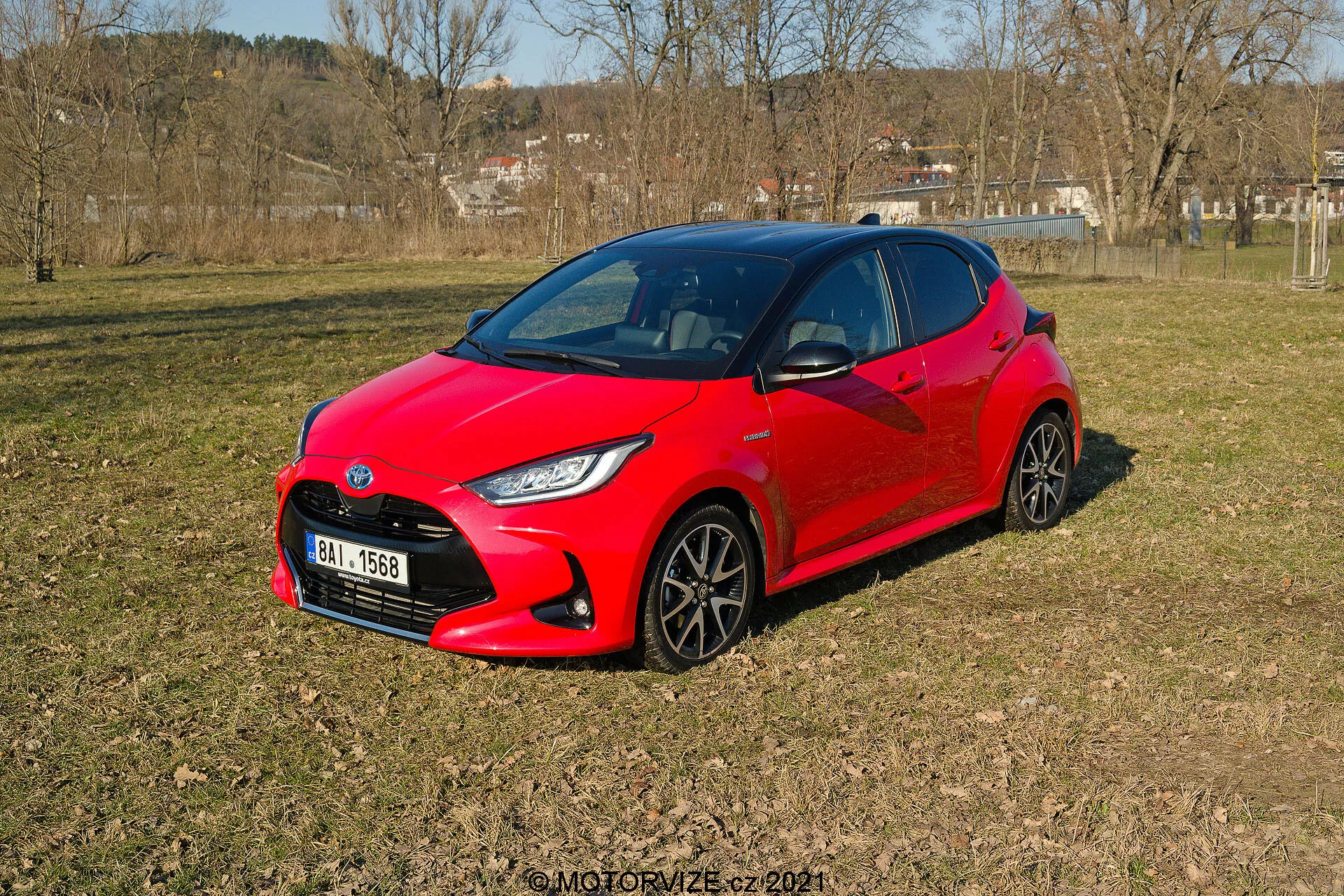 A imagem mostra um vibrante Toyota Yaris 2020 vermelho, repousando sobre um terreno gramado e irregular em uma área semi-rural, visto de uma perspectiva de três quartos. O design moderno e aerodinâmico do carro apresenta ângulos acentuados ao redor dos faróis e uma aparência compacta e esportiva. Rodas escuras de múltiplos raios e faróis LED estilizados são visíveis. As janelas laterais e o para-brisa dianteiro oferecem uma visão escura do interior, sugerindo possíveis sistemas automotivos avançados.