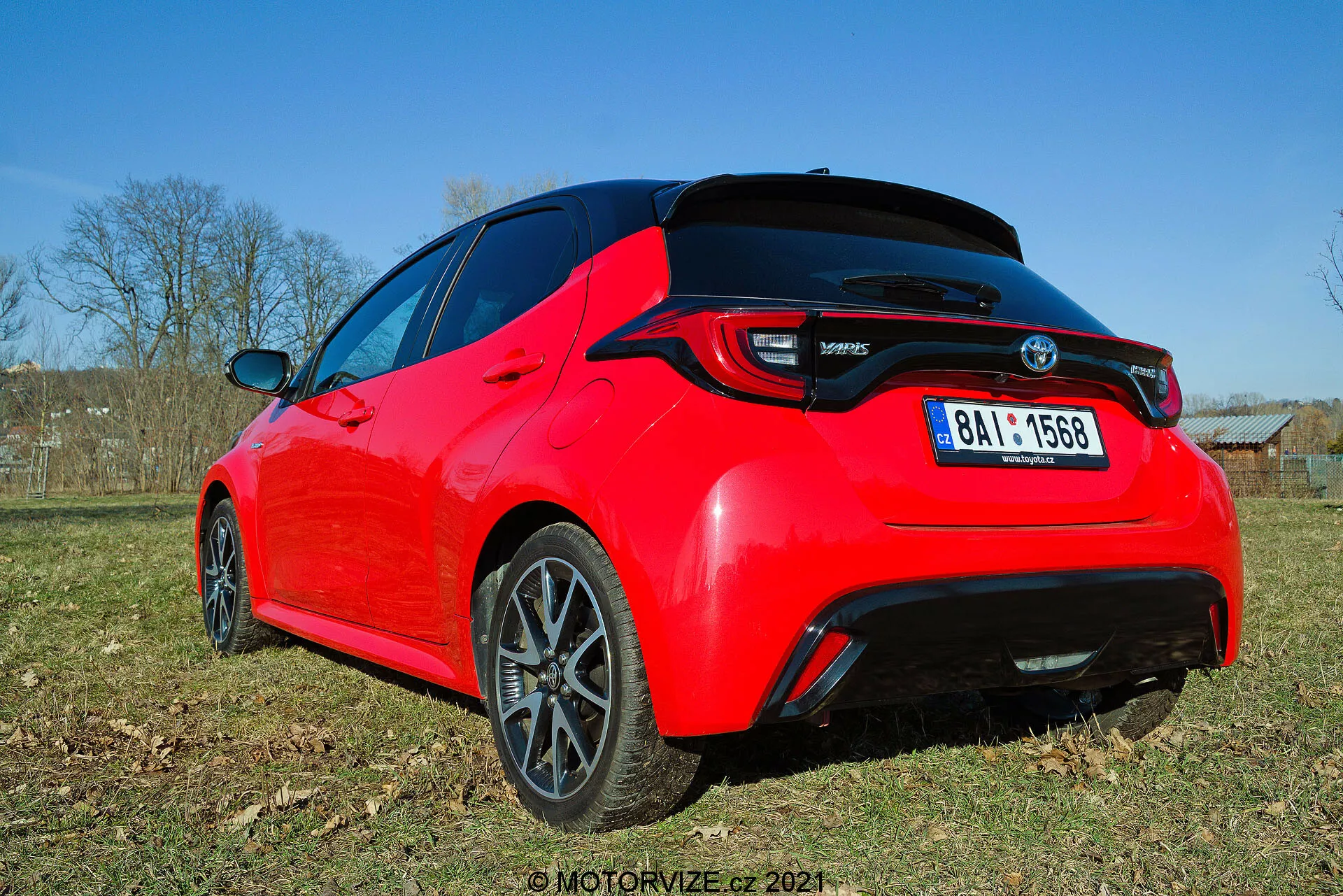 Vista traseira de três quartos de um hatchback Toyota Yaris 2020 vermelho brilhante, vista de um ângulo baixo, destacando linhas de design e características chave como luzes traseiras LED, spoiler traseiro, antena tipo barbatana de tubarão e rodas de liga leve com acabamento escuro. A foto captura o detalhe da traseira e parte do perfil lateral à luz do dia, mostrando sua estética esportiva e elementos modernos.