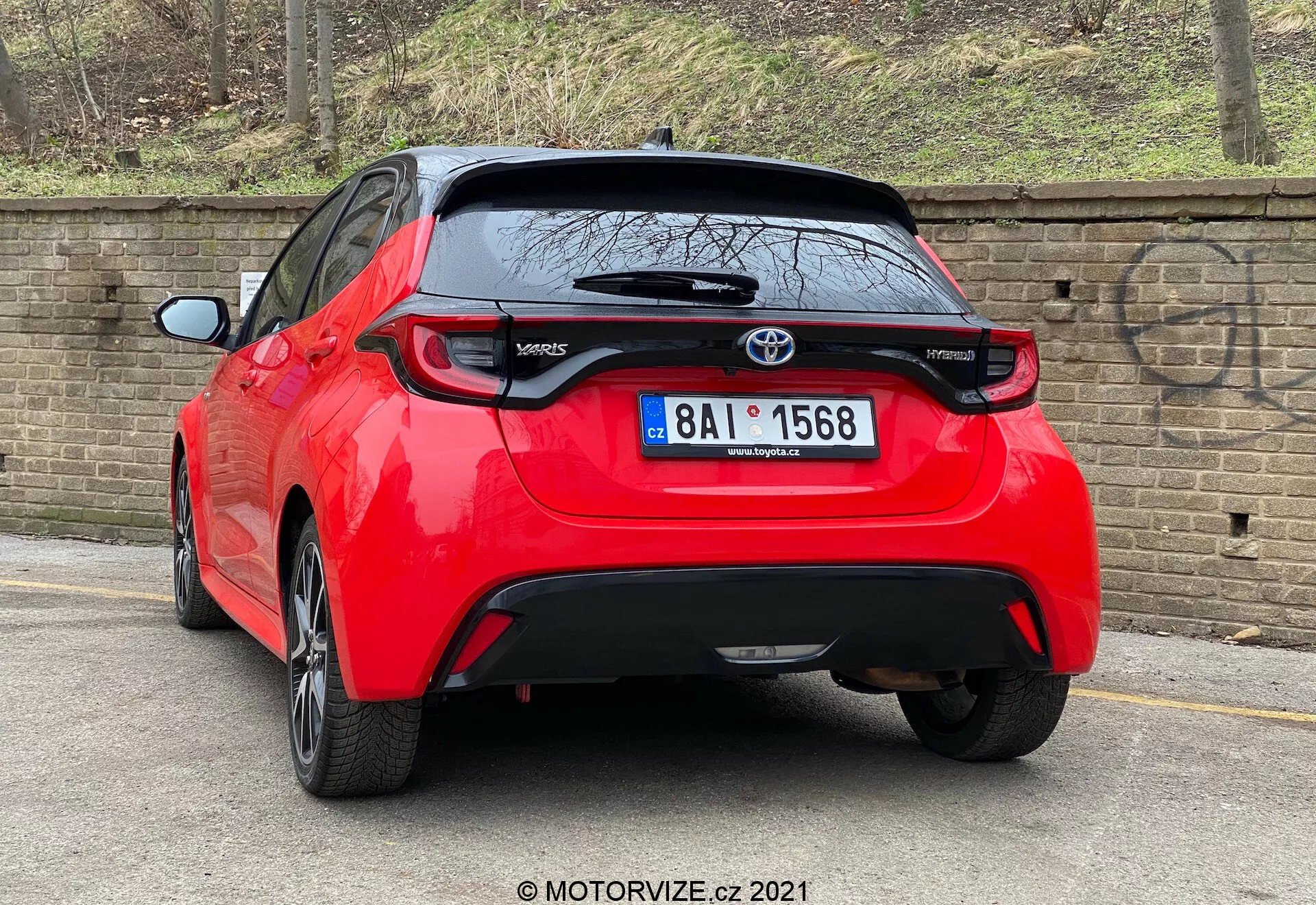 Uma imagem de alta resolução de um Toyota Yaris 2020 hatchback vermelho visto de um ângulo traseiro de três quartos. O veículo apresenta luzes traseiras distintas, um emblema Toyota centralmente colocado acima de uma provável câmera de ré e o emblema do modelo 'Yaris' na parte inferior esquerda da porta do porta-malas. Abaixo da área da placa de licença há uma peça de acabamento preta que pode ser de plástico ou fibra de carbono, com um pequeno spoiler traseiro que se estende sobre a janela traseira. O carro, estacionado perto do meio-fio em uma rua pavimentada ao lado de um muro de tijolos, sugere um ambiente urbano ou residencial.