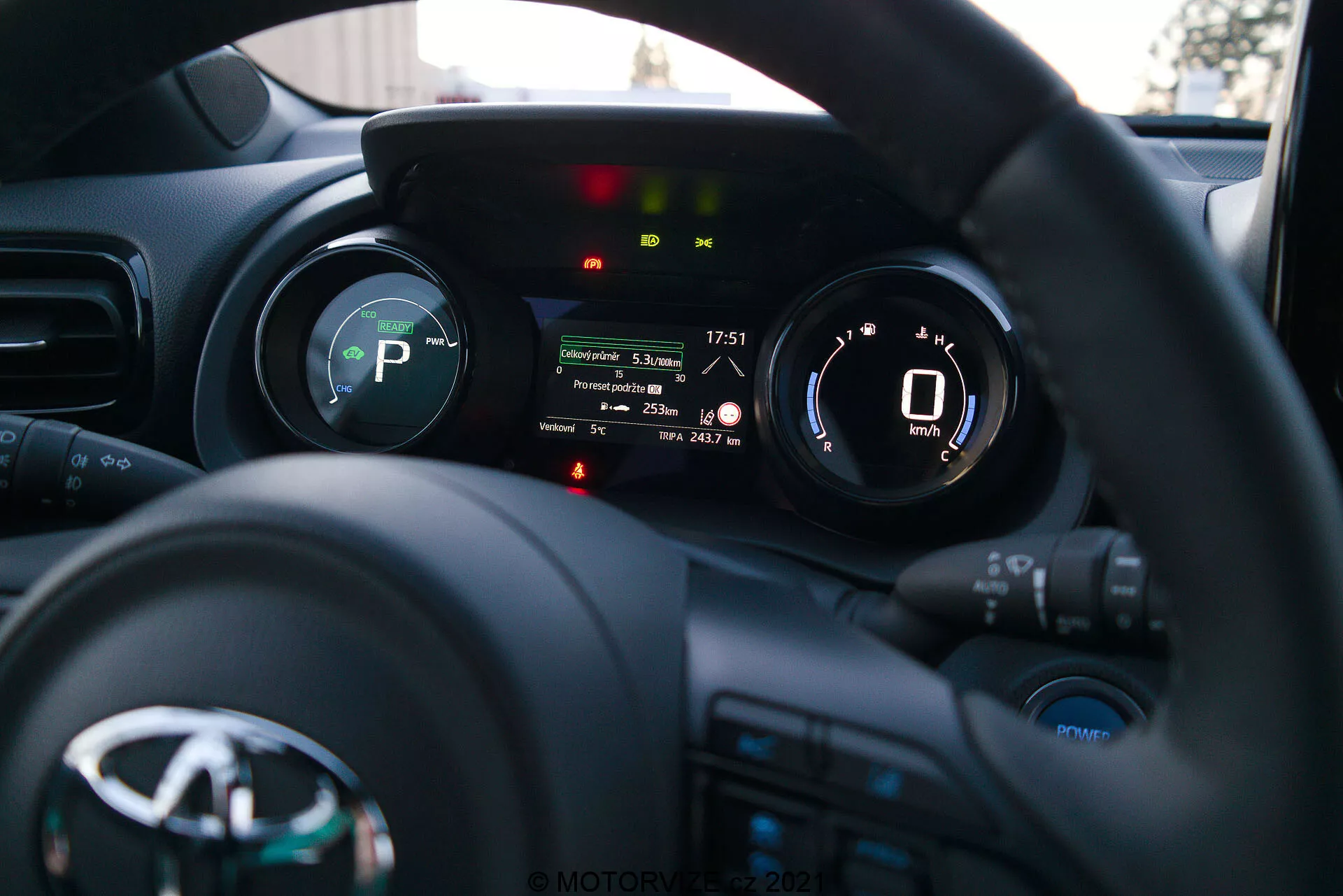 Uma fotografia do interior do Toyota Yaris 2020 da perspetiva do condutor, focando o volante e o painel de instrumentos. O volante, com o logotipo do fabricante e vários botões de controlo para mídia, comunicação e controlo de cruzeiro, está na parte inferior. O painel de instrumentos atrás do volante possui um visor digital que mostra um velocímetro a 0 km/h e indicadores para a marcha (P de Park), lembrete do cinto de segurança, alerta de porta aberta e outras luzes de status do veículo. A baixa profundidade de campo destaca o painel de instrumentos, enquanto o painel e o interior do carro estão suavemente desfocados no fundo. A imagem, provavelmente tirada no final da tarde ou início da noite, mostra instrumentação moderna e tecnologia de interface de veículos.