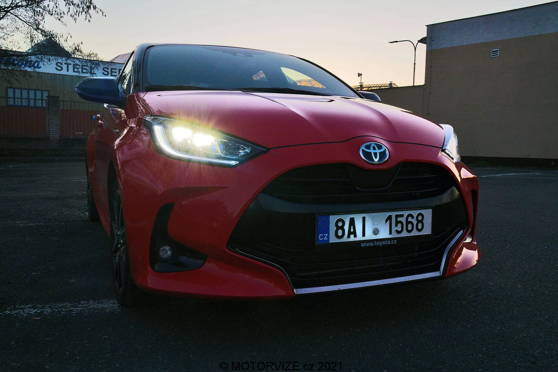 Close-up em ângulo baixo de um Toyota Yaris 2020 vermelho estacionado ao ar livre ao nascer ou pôr do sol. Grade frontal, para-choques, capô esculpido aerodinamicamente, faróis em LED afilados e grandes entradas de ar destacando o emblema da Toyota. Placa europeia visível, situada contra um fundo industrial ou comercial com prédio 'STEEL SERVICE'. Céu claro com luz em degradê destacando o design esportivo do veículo.